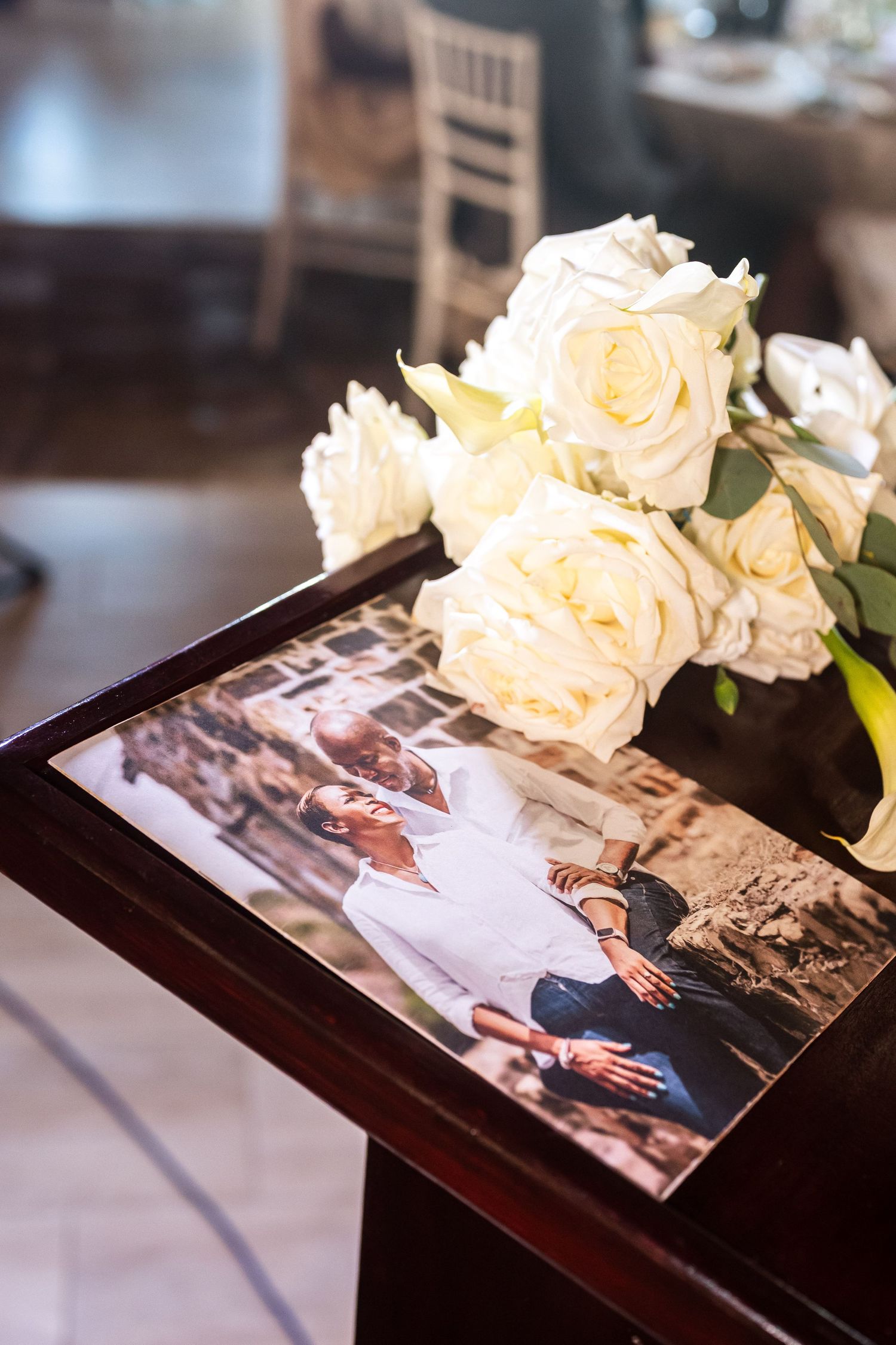 White roses bouquet resting on a wooden table beside a couple's photo print at a wedding reception venue.