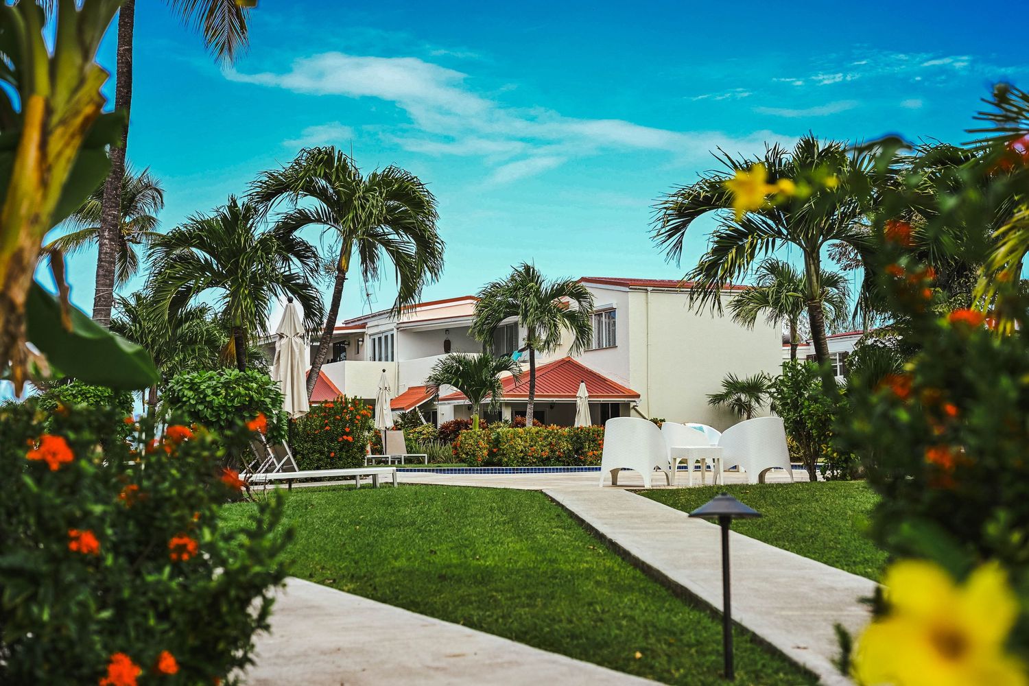 Lush tropical resort grounds with palm trees, colorful flowers, manicured lawn, and white buildings.