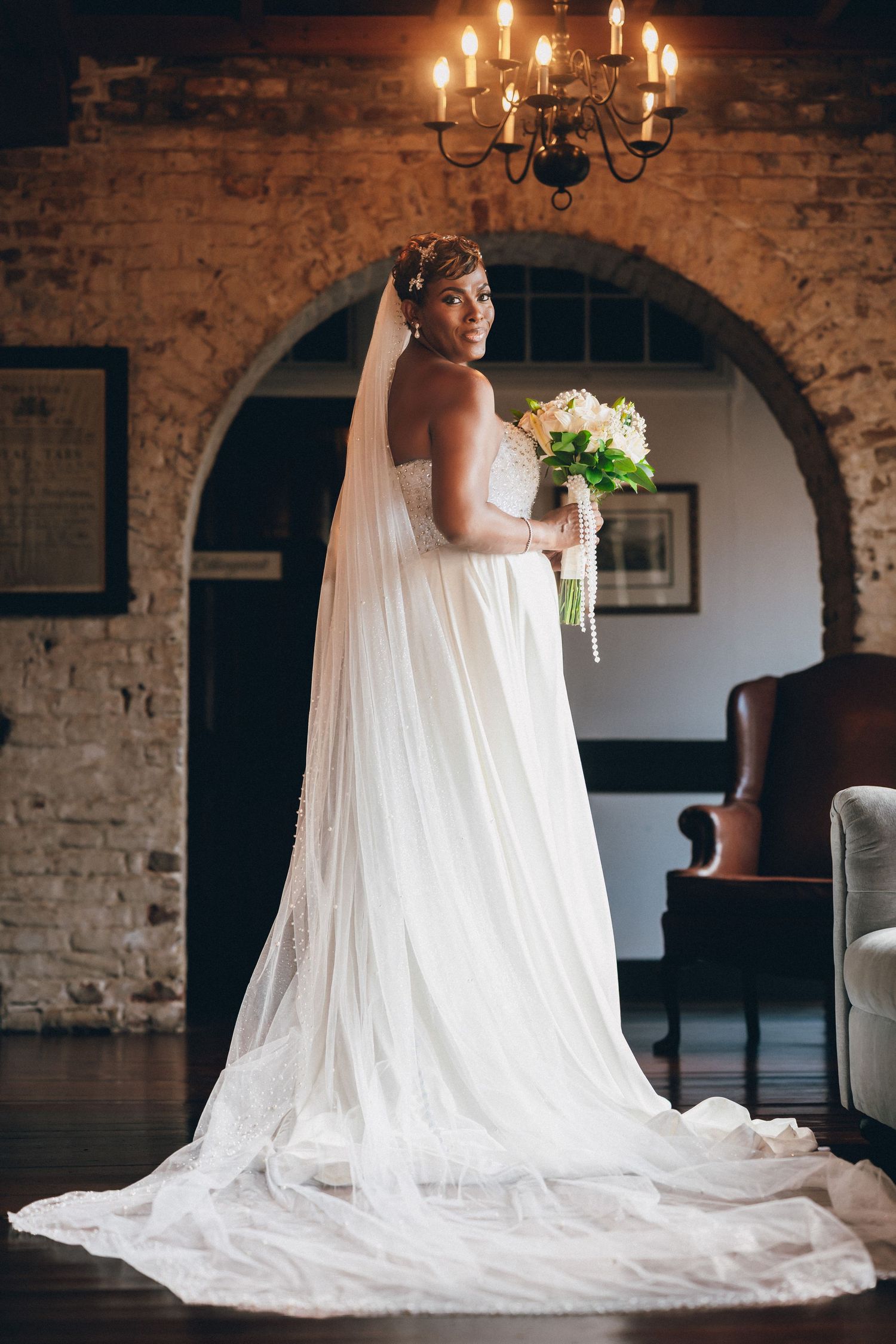 Bride in white strapless gown with long veil holds bouquet under chandelier in rustic brick archway venue.