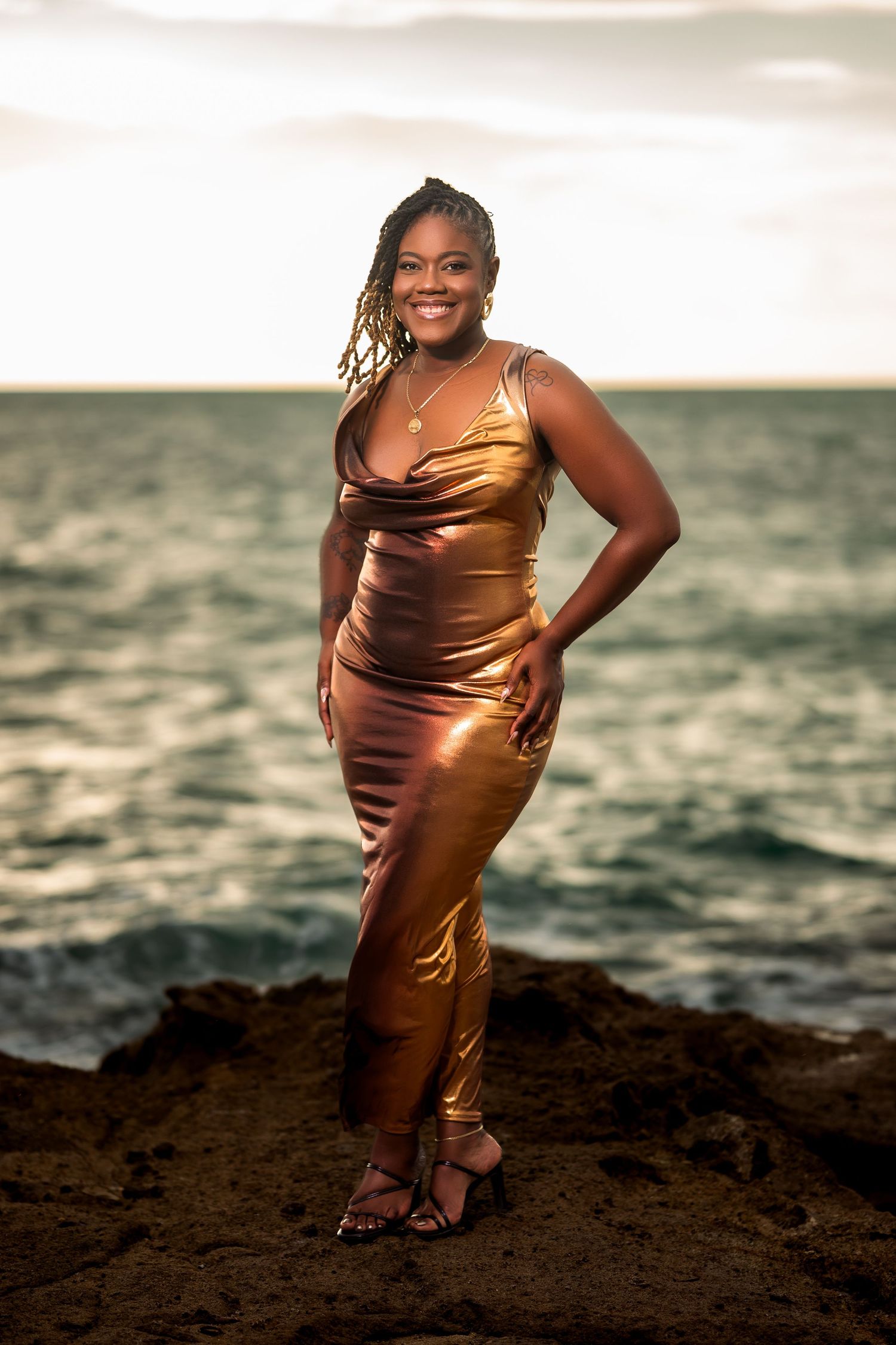 Woman in gold satin gown posing on rocky shore at sunset with ocean waves in background.