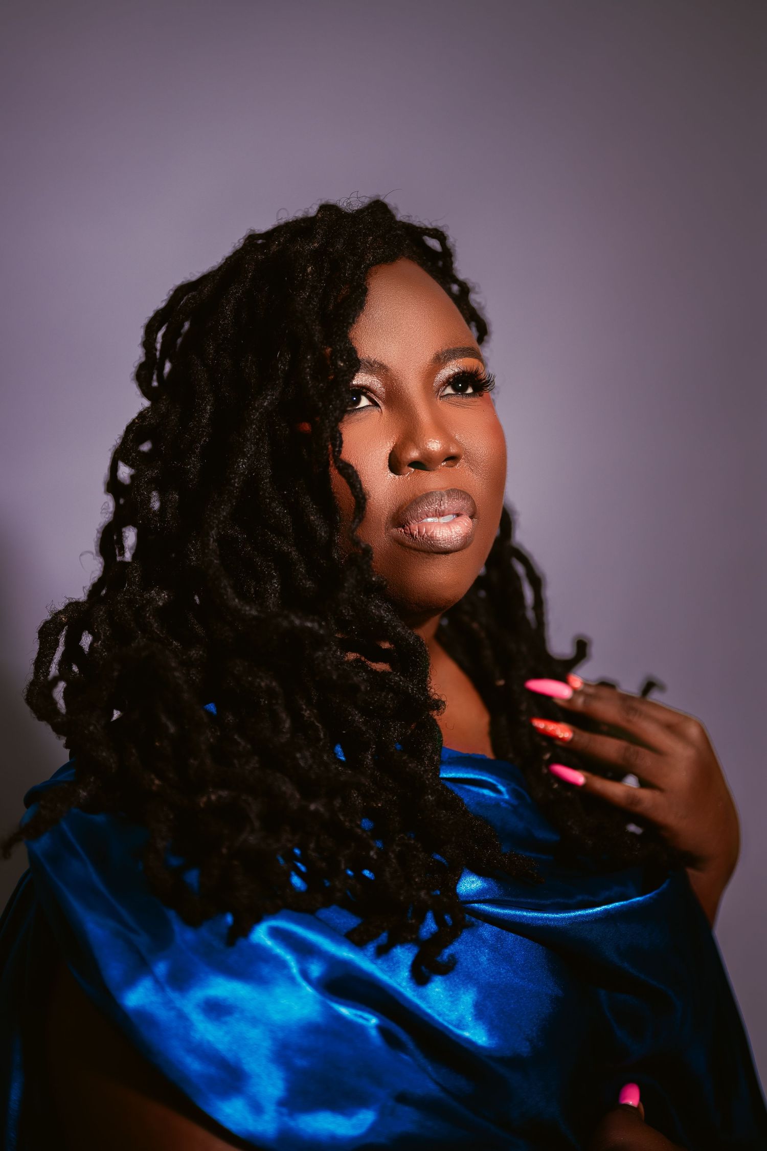 A Black woman with long locs wearing a blue satin wrap gazes upward with pink nails resting on her shoulder.