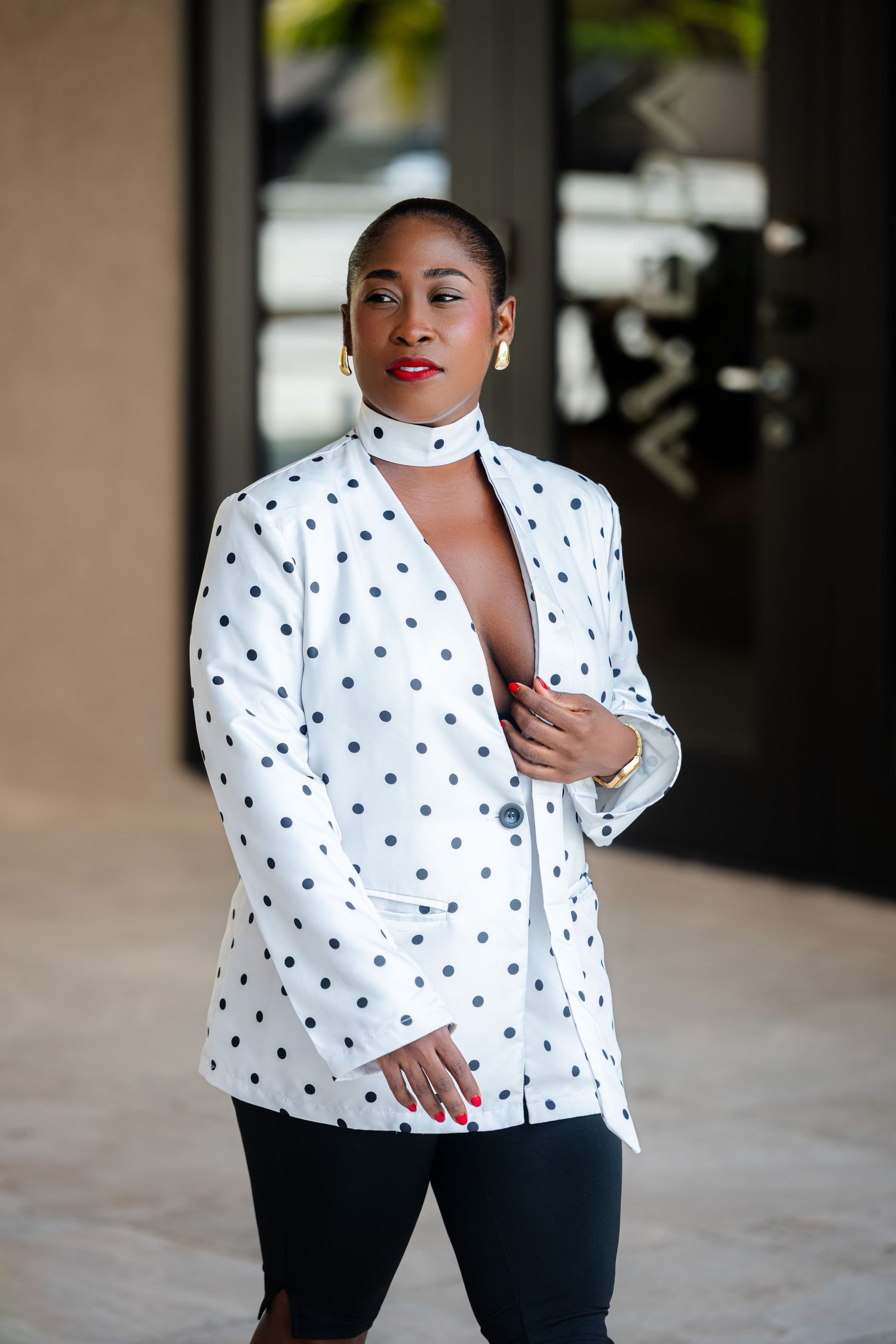 Stylish Black woman in white polka dot blazer with gold earrings and red nails posing near glass doors.