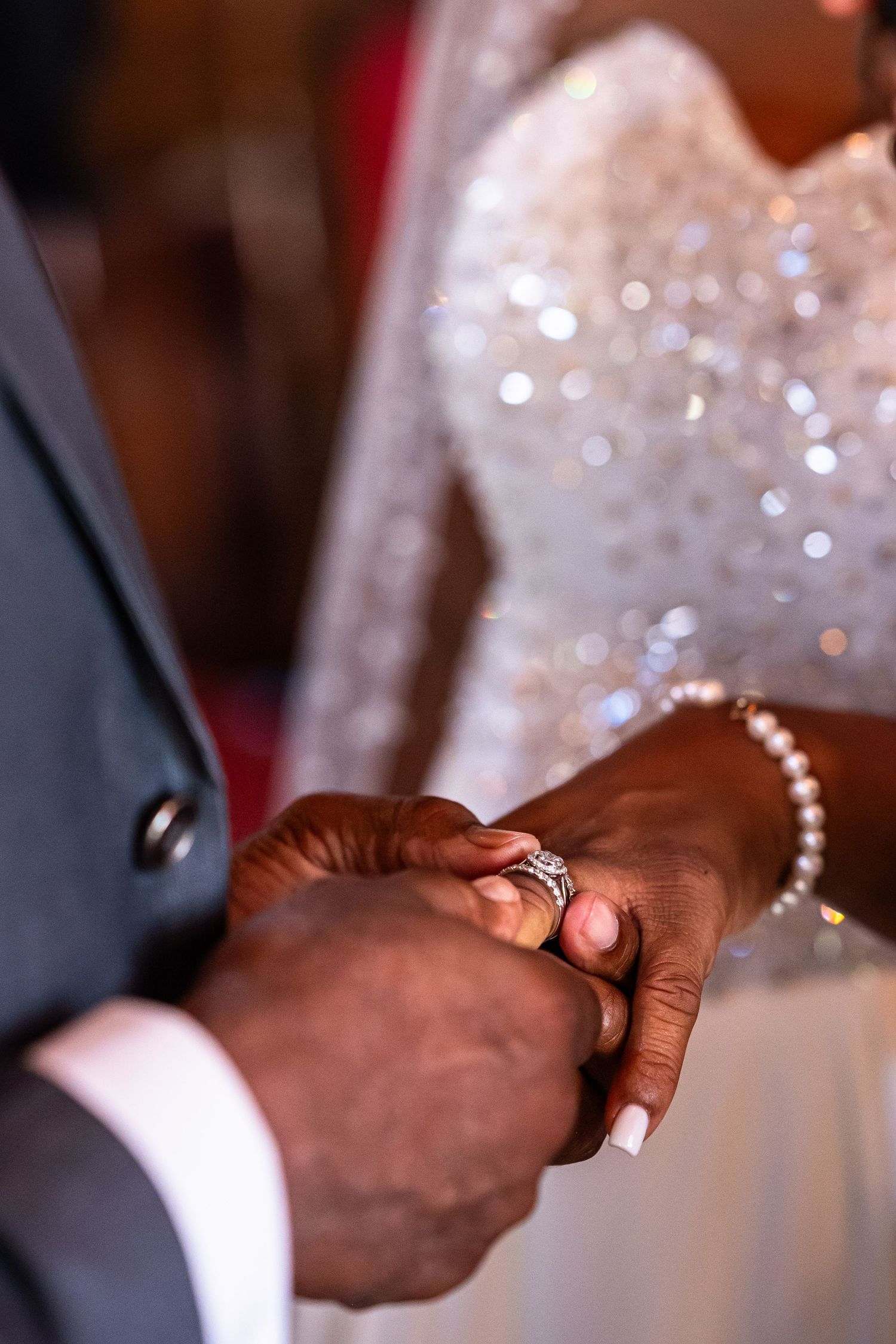A groom places a diamond ring on his bride's hand during a wedding ceremony, with her pearl bracelet visible.