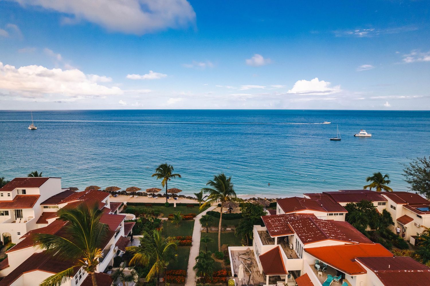 Elevated view of a beachfront Caribbean resort with red-roofed buildings, palm trees, and sailboats on blue sea.