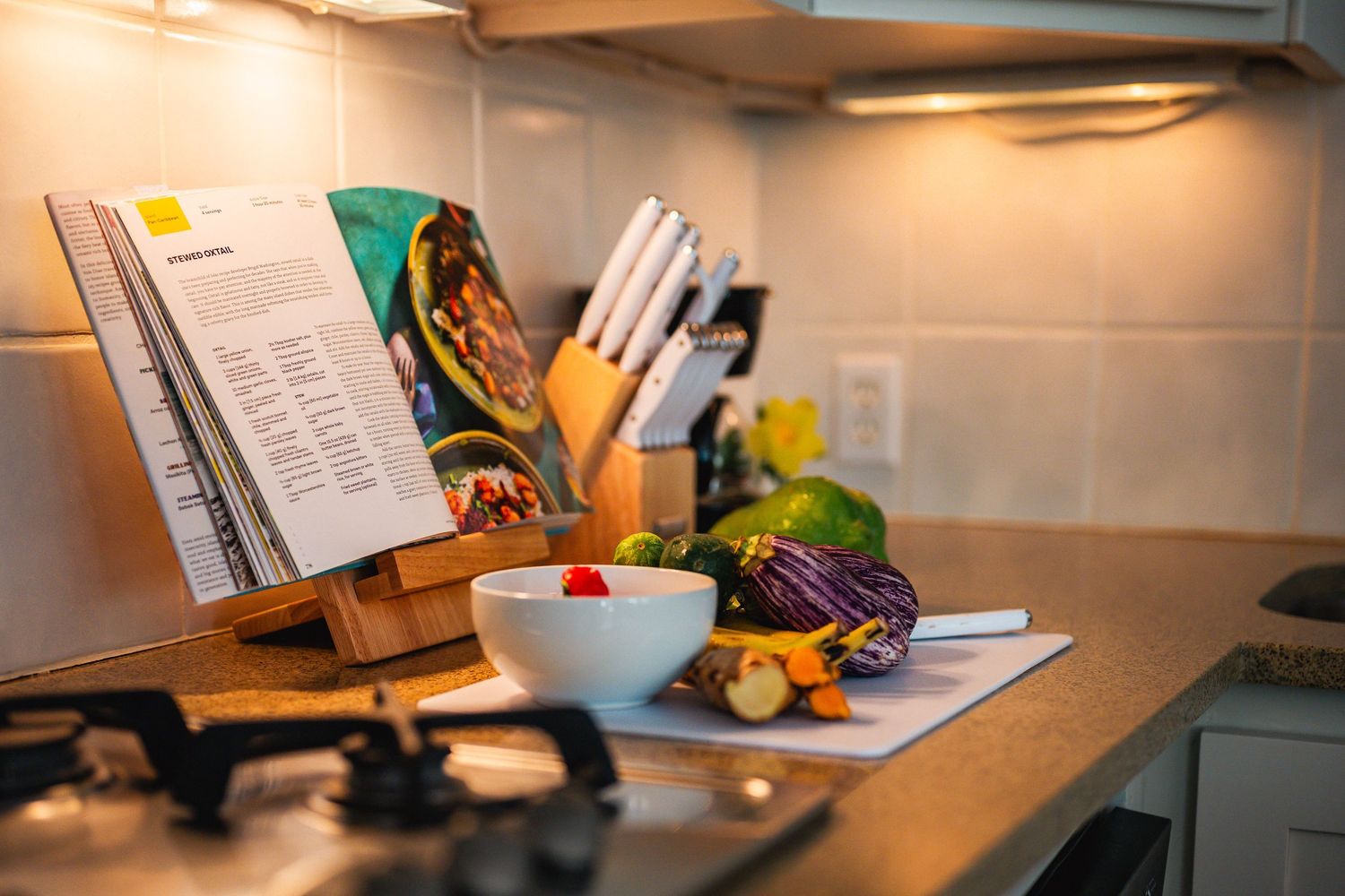 Kitchen counter with open cookbook, fresh vegetables, white bowl, and knife block under warm lighting.