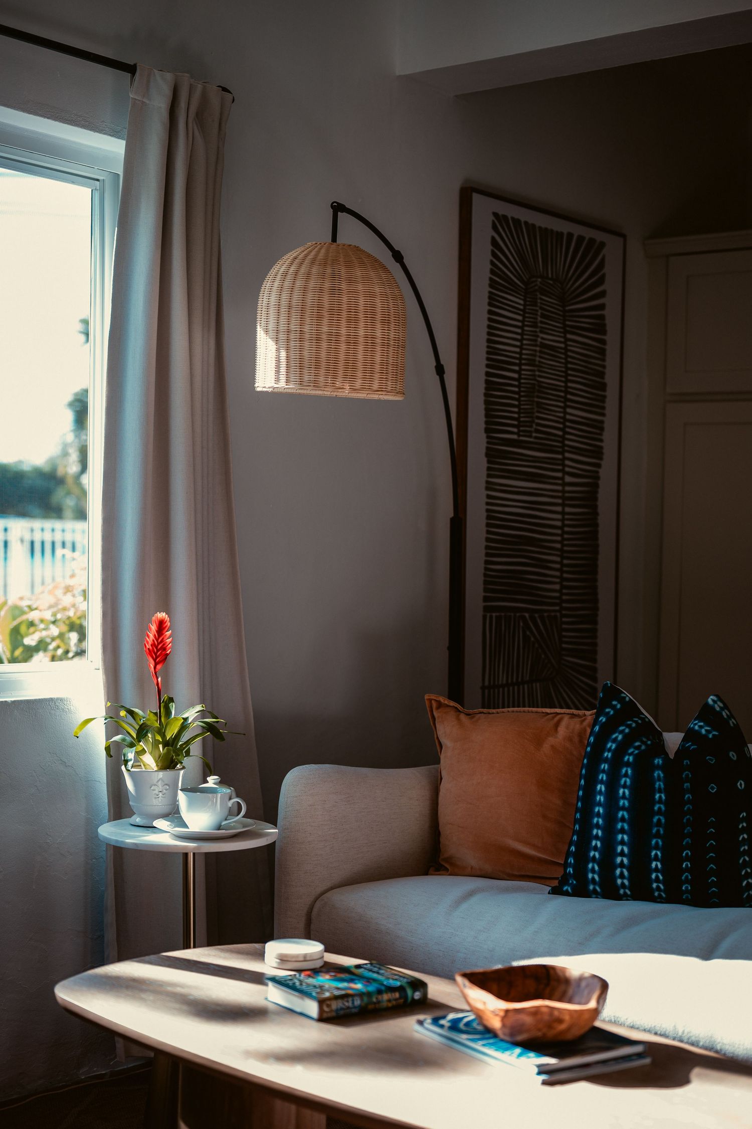 Cozy living room corner with rattan arc lamp, linen sofa, terracotta pillow, and sunlight through window.
