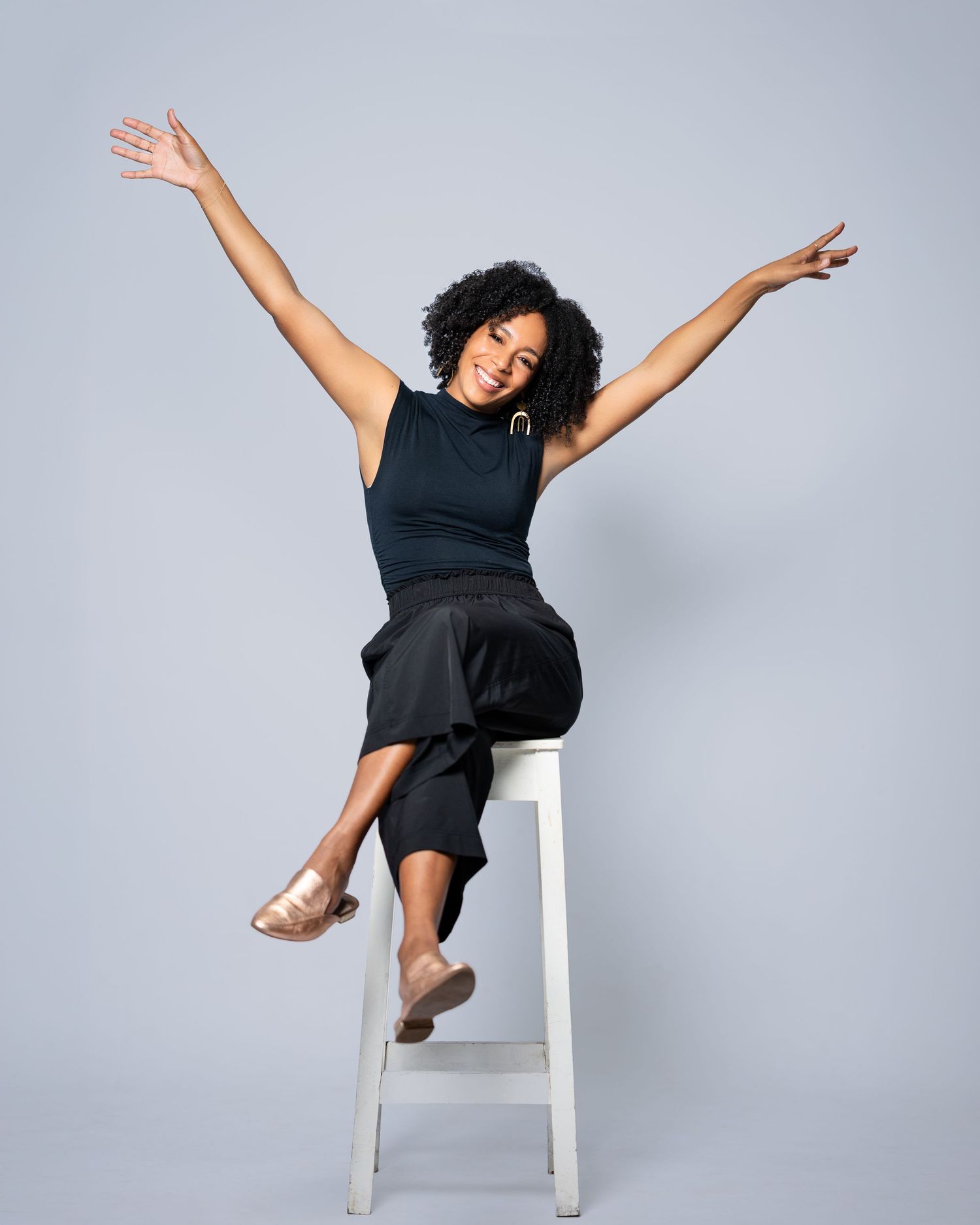 A joyful woman in black outfit sits on white stool with arms raised, wearing gold flats against gray background.