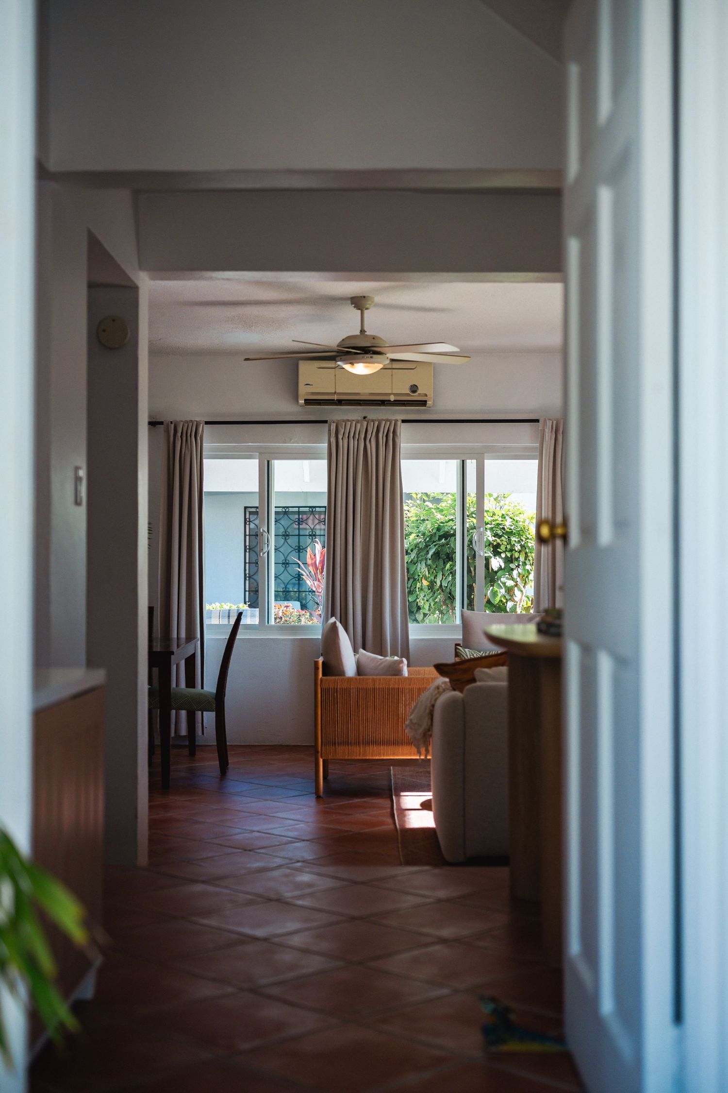 Hallway view into a bright tropical living room with terracotta floors, ceiling fan, and large windows.