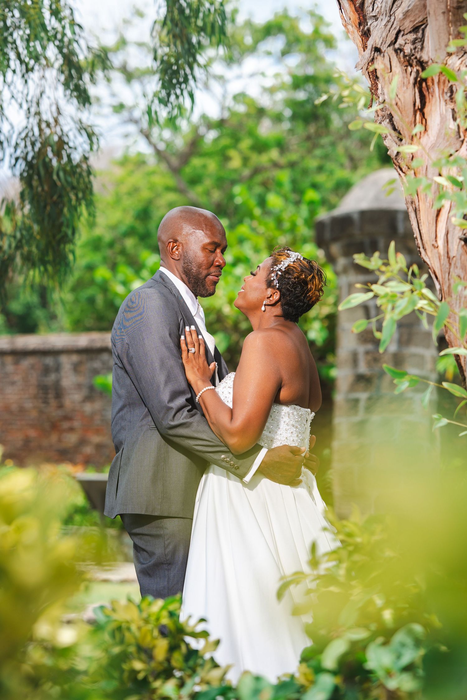 A bride and groom share a romantic moment in a lush garden, dressed in a white gown and gray suit.
