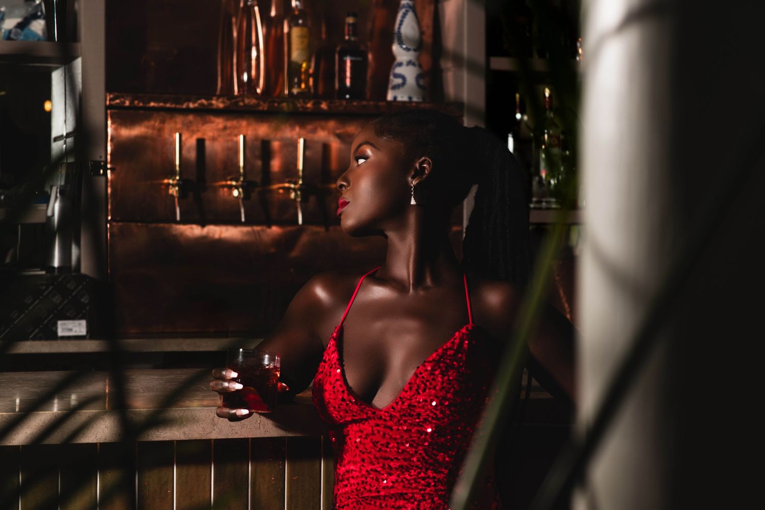 A woman in a red sequin dress holds a wine glass at an upscale bar, gazing upward in ambient candlelight.
