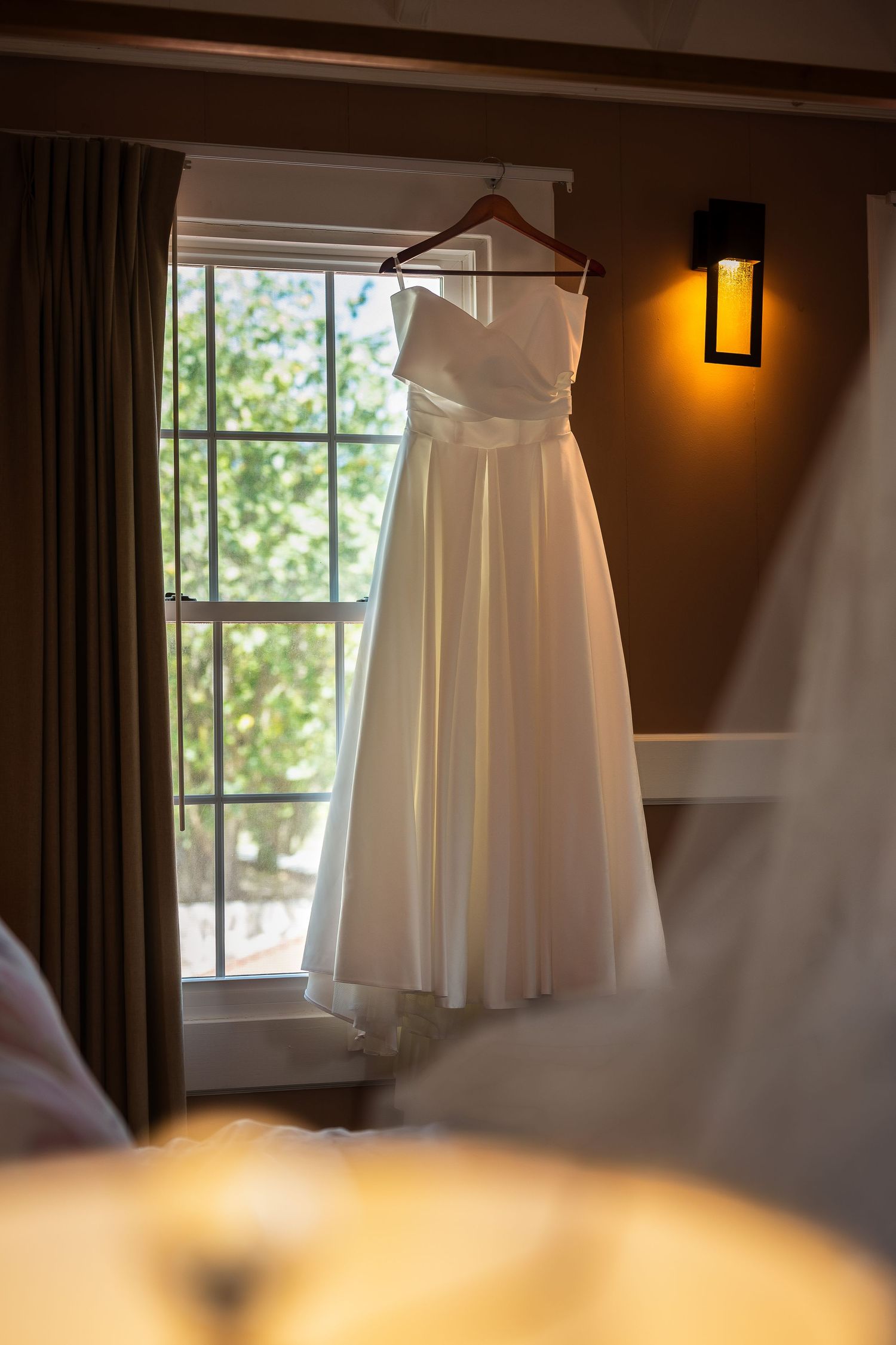 White strapless wedding dress hanging on wooden hanger in front of window with warm wall sconce lighting.