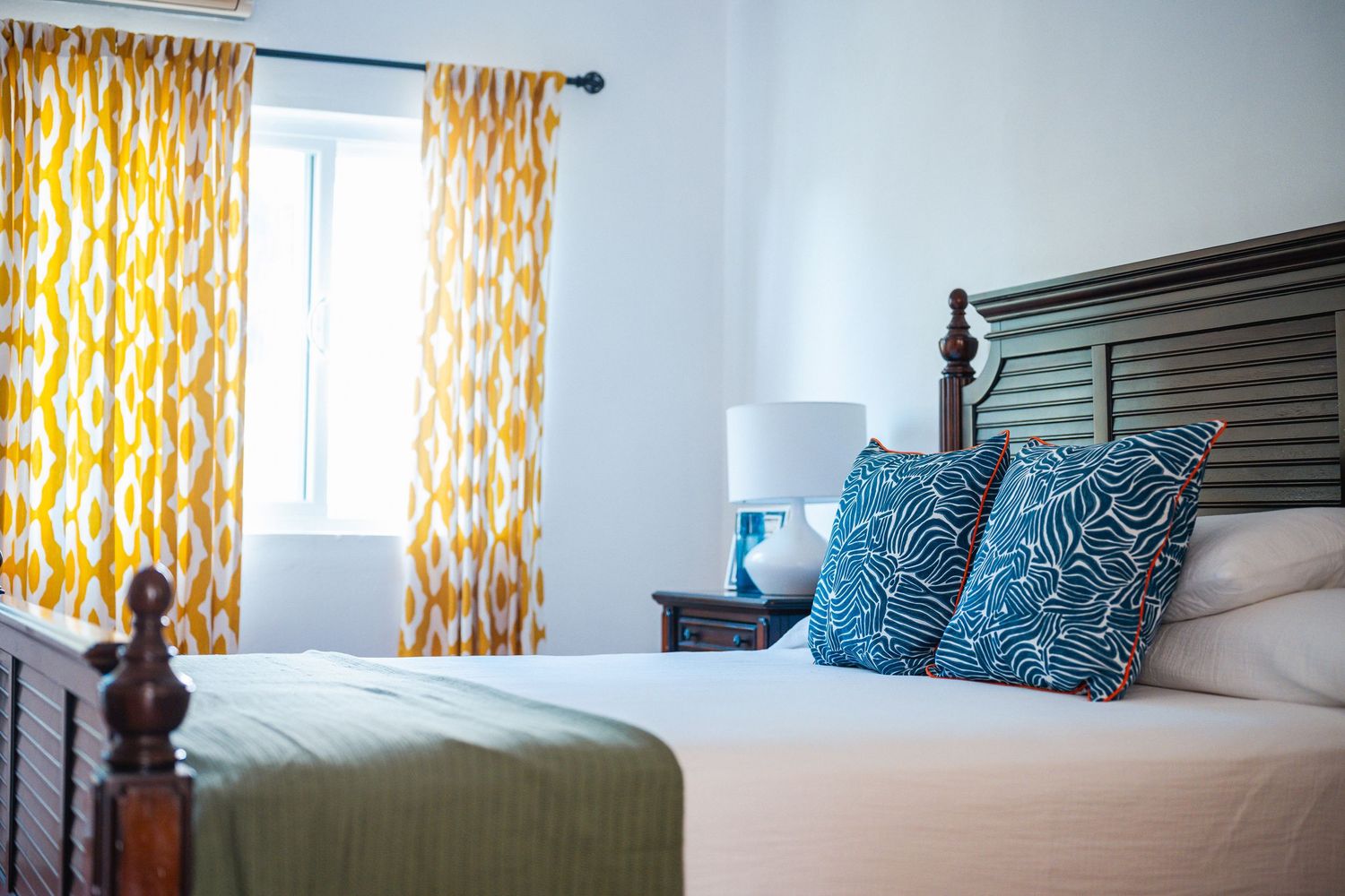 Elegant bedroom with dark wood headboard, blue leaf-print pillows, green bedspread, and vibrant yellow patterned curtains.