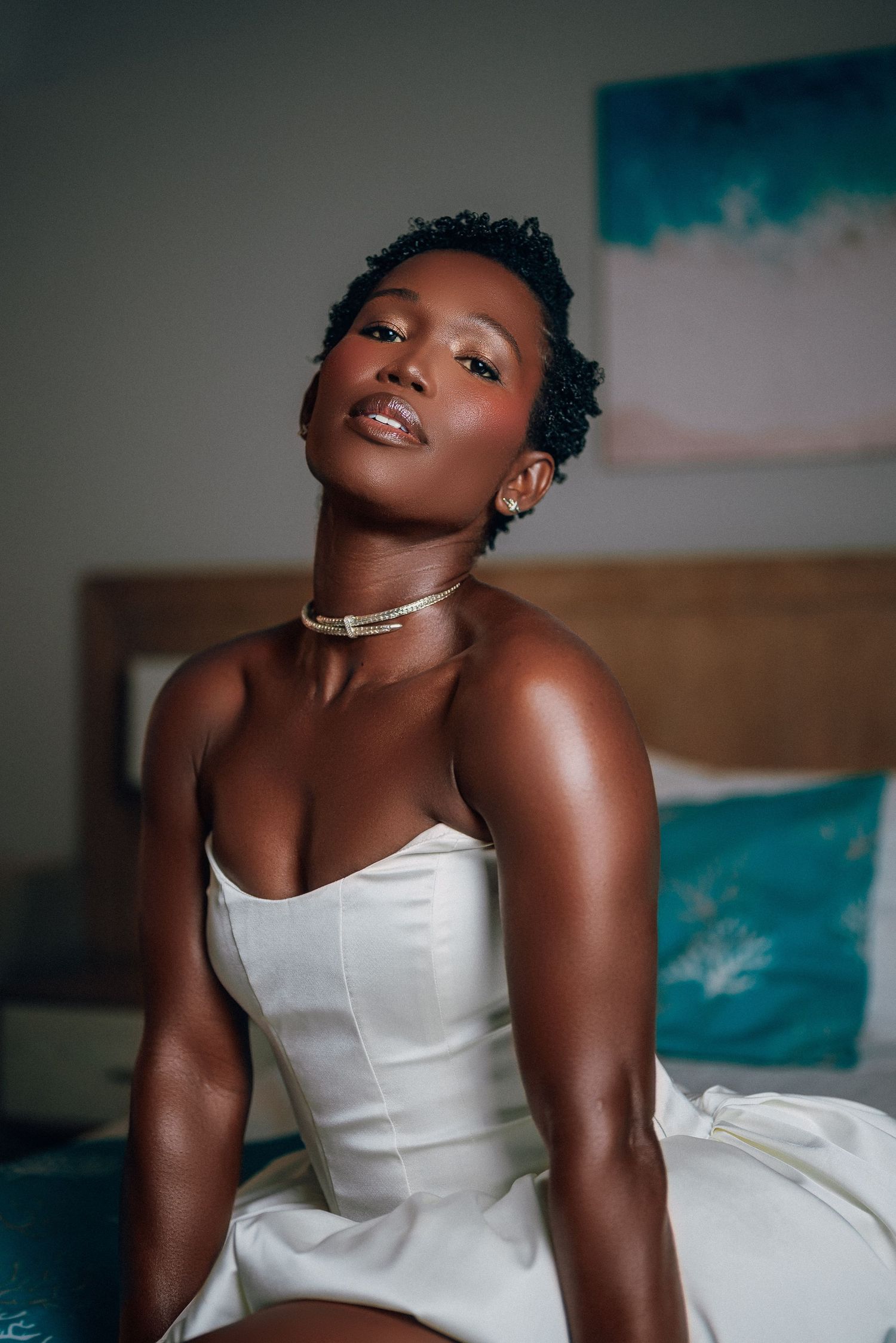Beautiful Black bride in strapless white dress with gold choker necklace sitting on bed in hotel room.