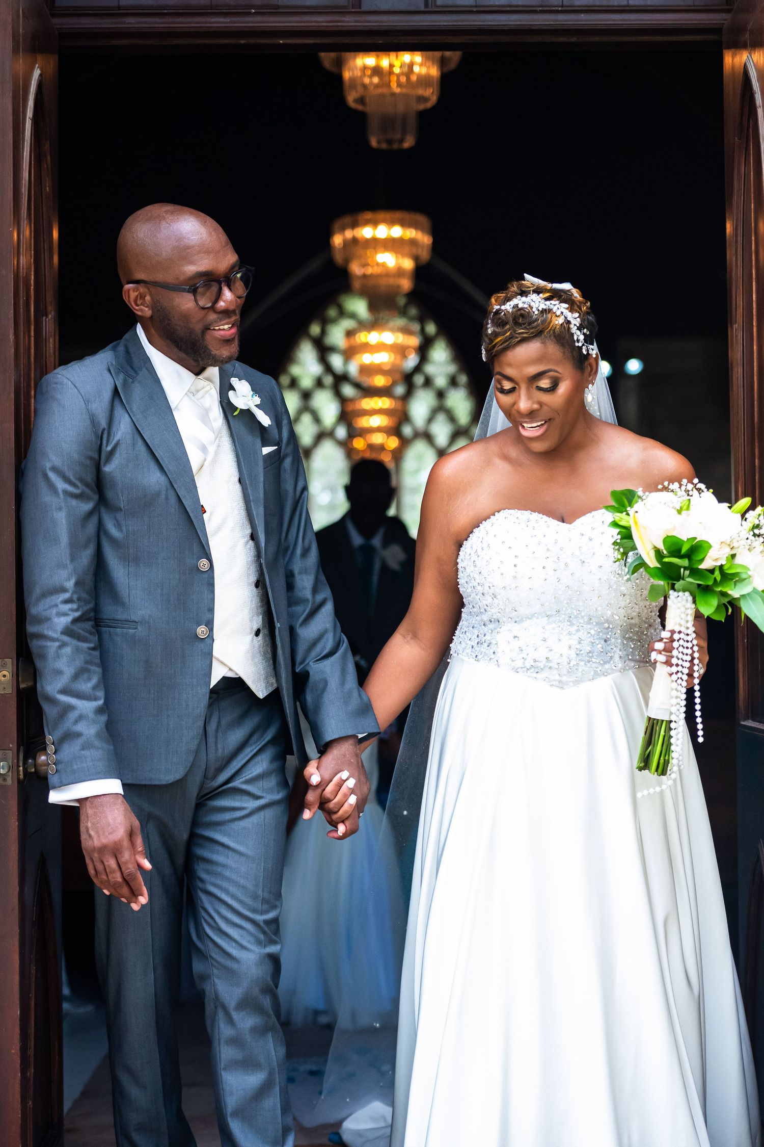 Newlywed couple holding hands exiting church, groom in gray suit, bride in strapless beaded gown holding white bouquet.