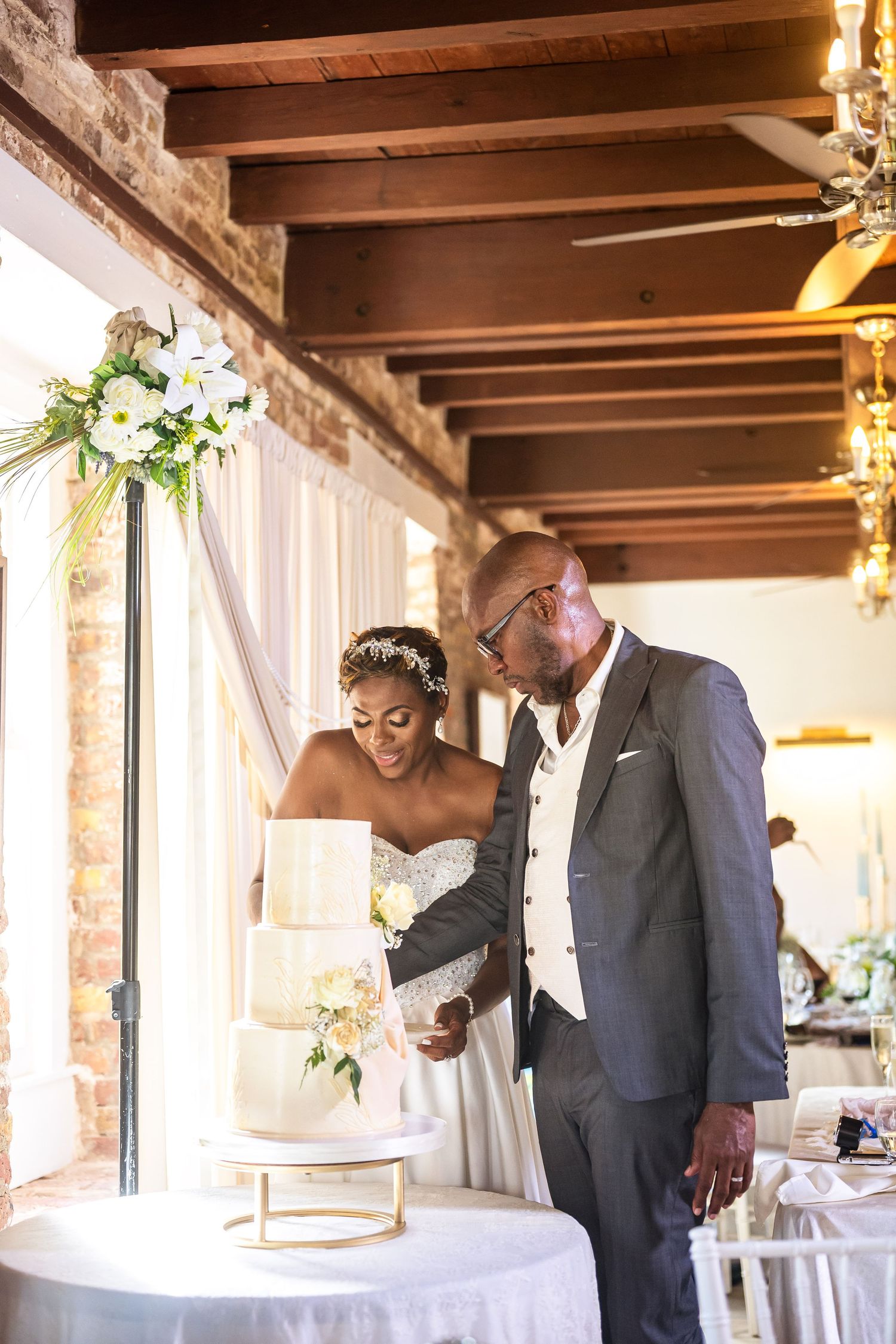 Black couple cutting white wedding cake at elegant reception venue with exposed brick walls and floral arrangements.