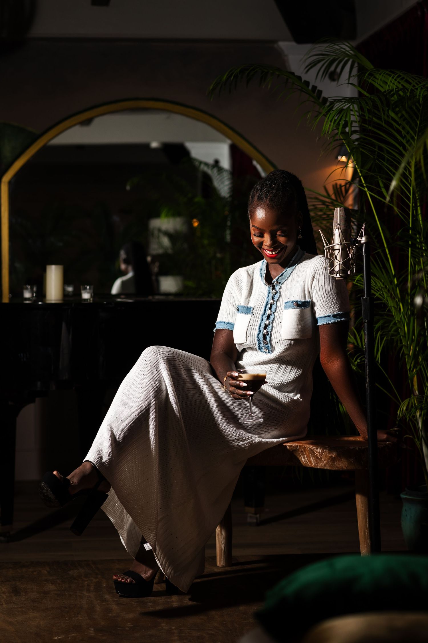 Smiling woman in white dress sitting on bench, using smartphone in dimly lit restaurant with tropical plants.
