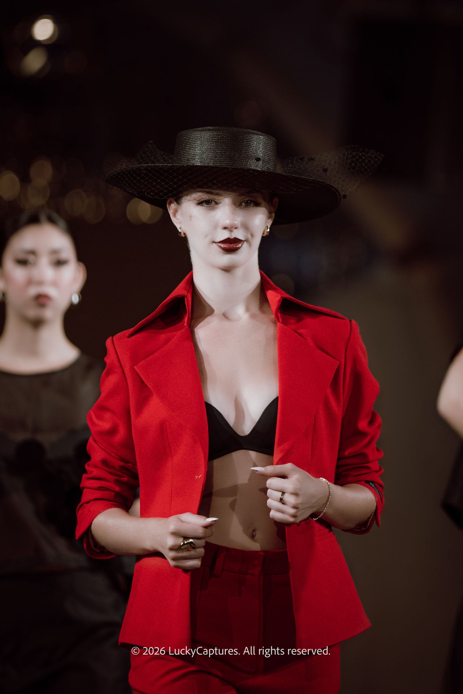 Model in red blazer and black wide-brim hat with dark lipstick walks runway at fashion show.