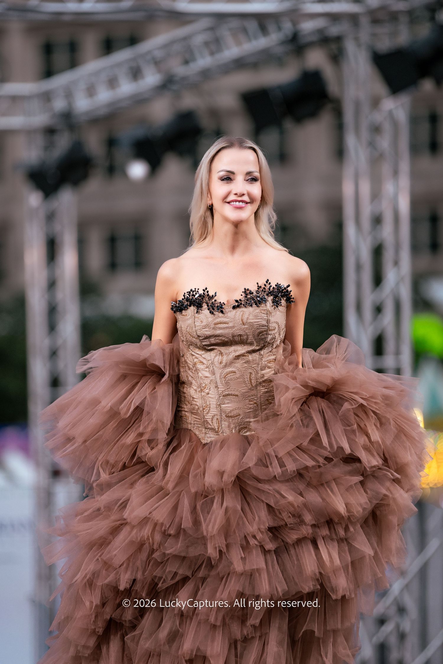 Model poses in brown ruffle tulle gown with black floral lace strapless bodice on runway.