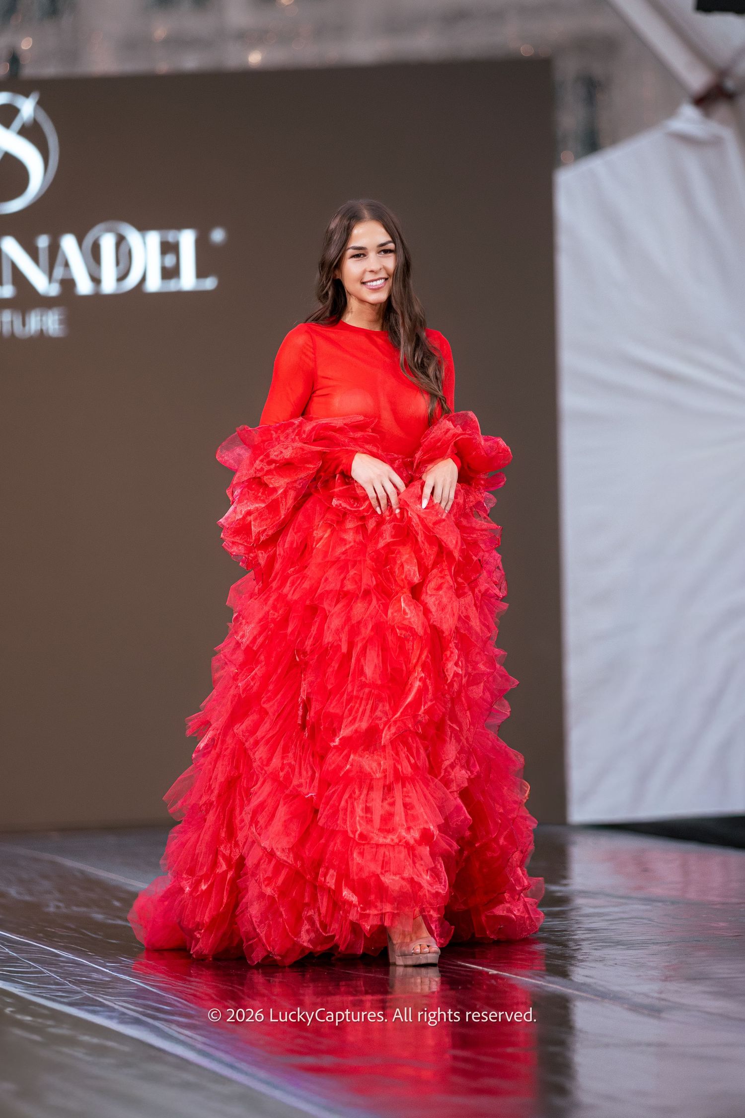 Model walks runway in a voluminous red ruffled tulle gown at a Nadel Couture fashion show.
