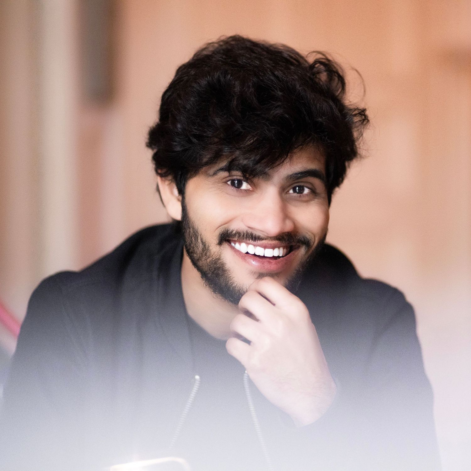 Young man with curly dark hair and beard smiling confidently, resting chin on hand against soft blurred background.