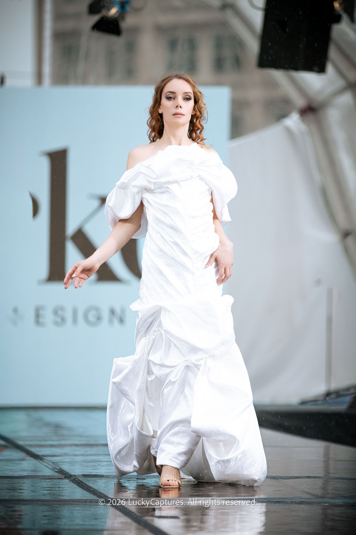 Model walks runway in elegant white off-shoulder ball gown at fashion show.