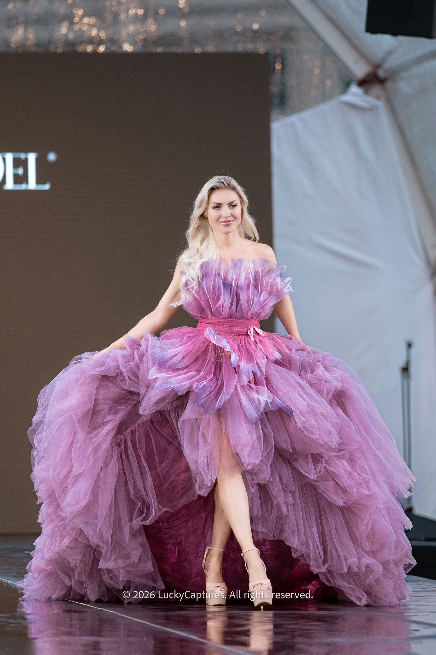 Blonde model walks in flowing pink ombre tiered tulle ball gown with high slit on wet outdoor runway.