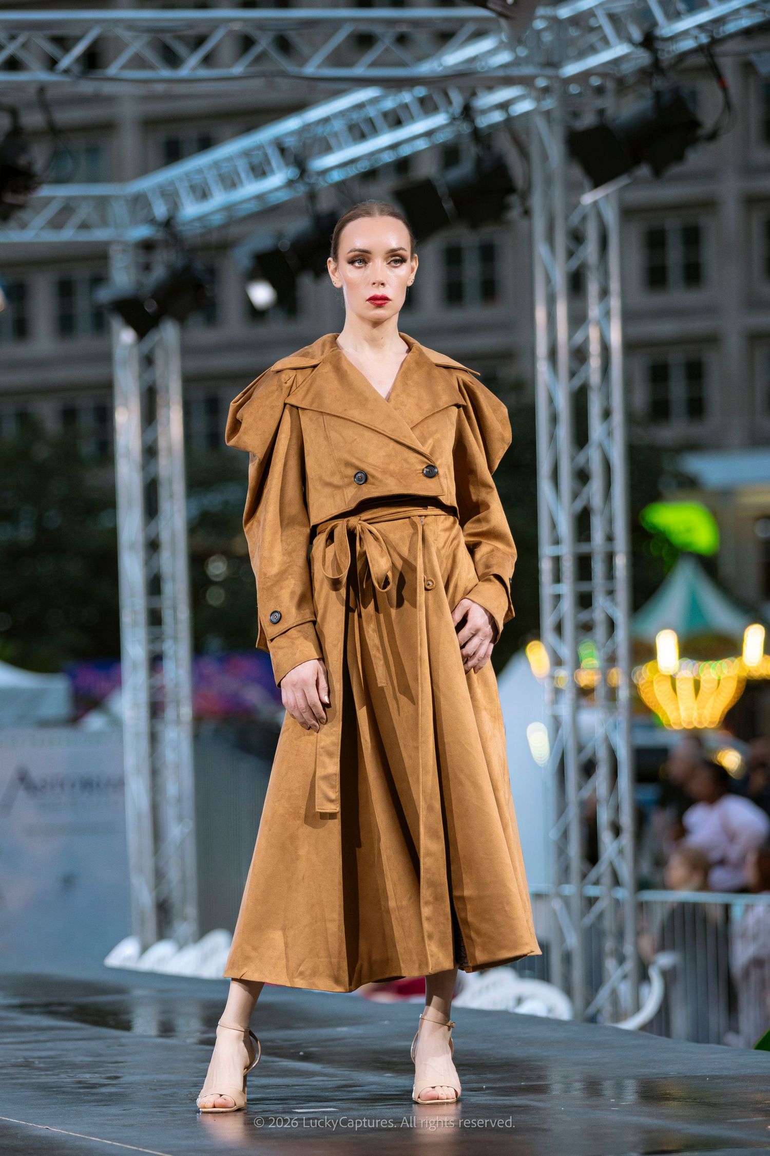 Model walks runway in elegant camel trench coat with puffed shoulders and belted waist at outdoor fashion show.