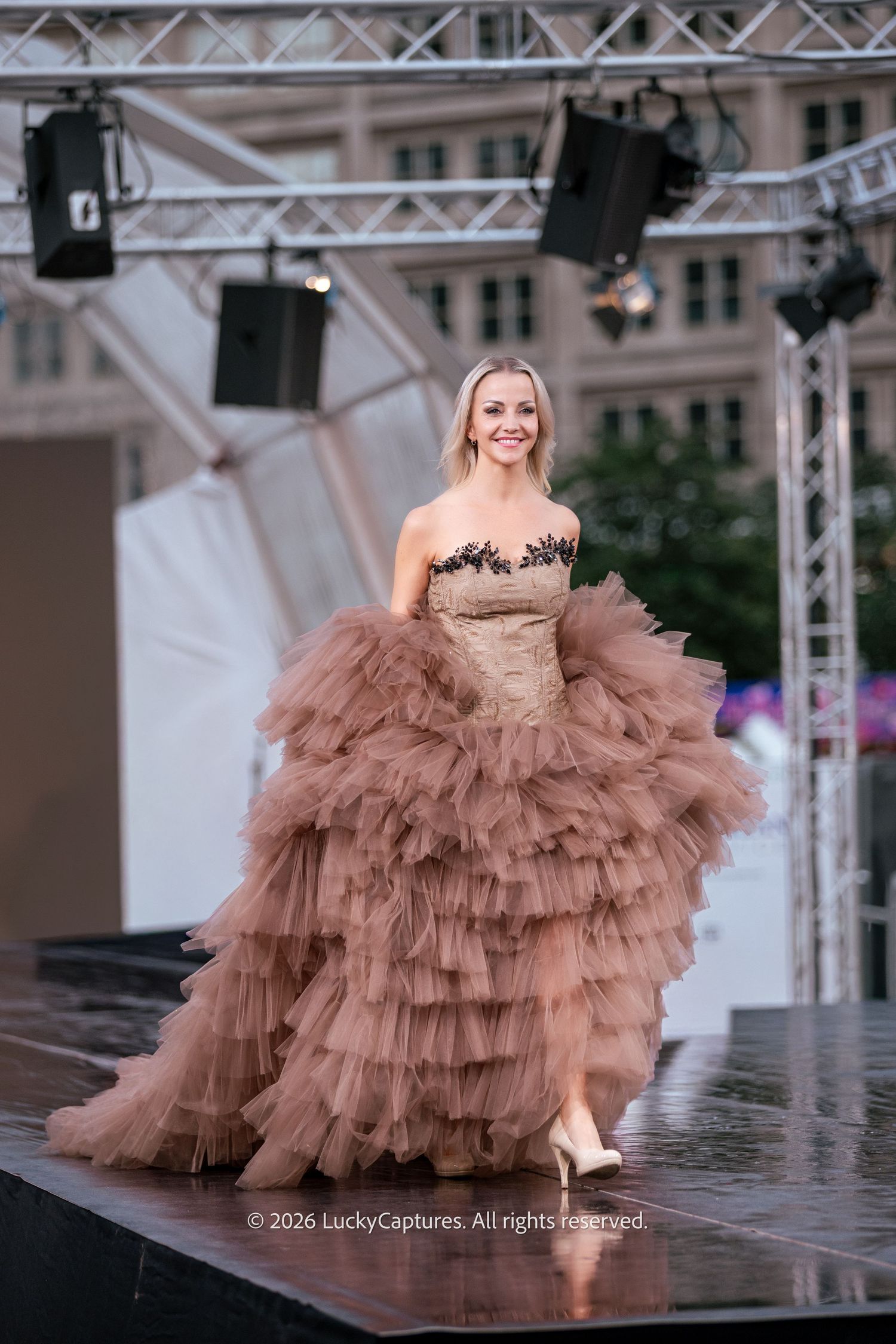 Model walks outdoor runway in voluminous brown tiered tulle ball gown with black lace bodice.