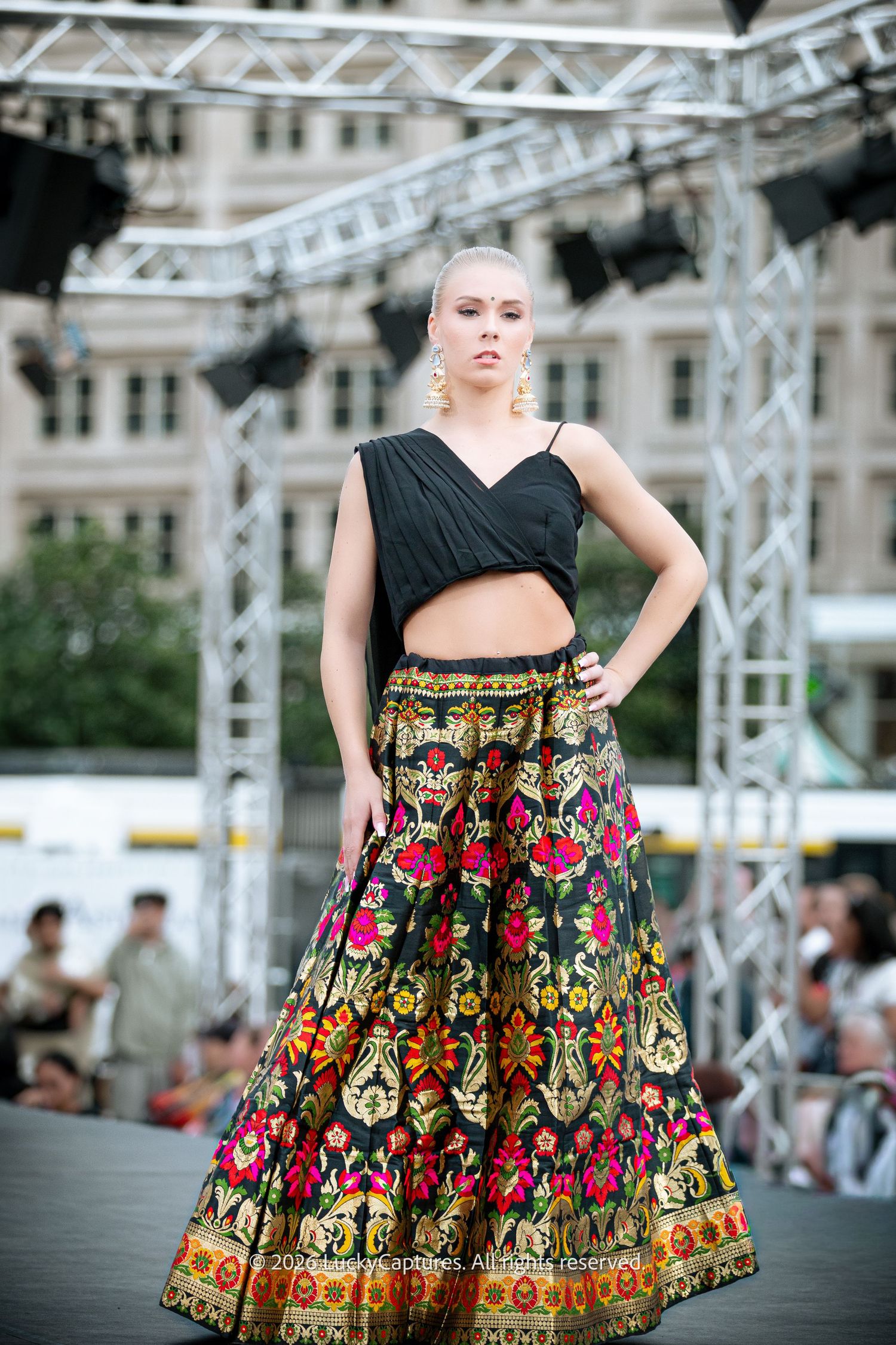 Model on runway wearing black crop top and vibrant floral embroidered skirt at outdoor fashion show.