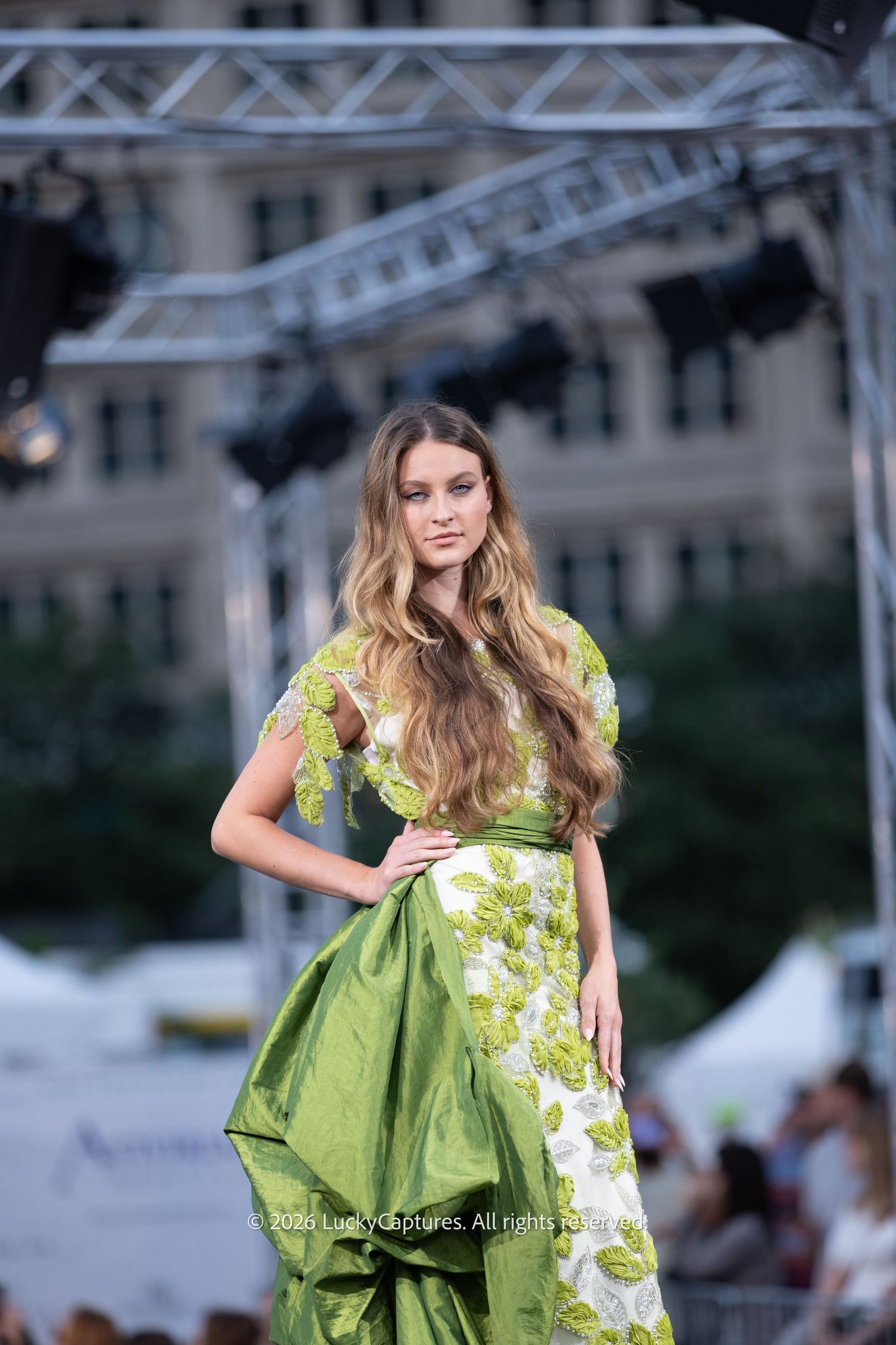 Model walks runway in elegant green floral gown at outdoor fashion show.