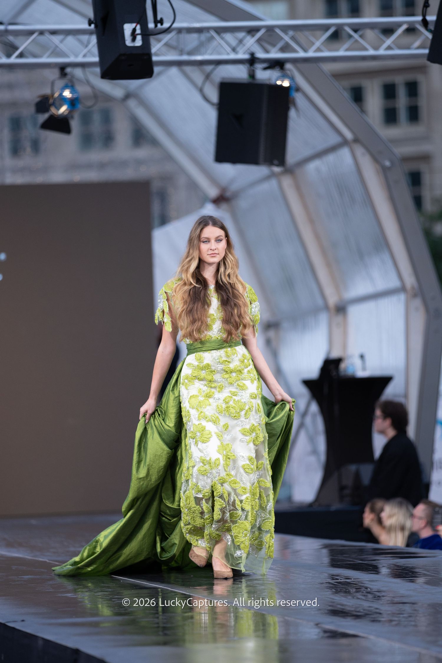 Model walks runway in elegant green floral gown with flowing silk train at outdoor fashion show.