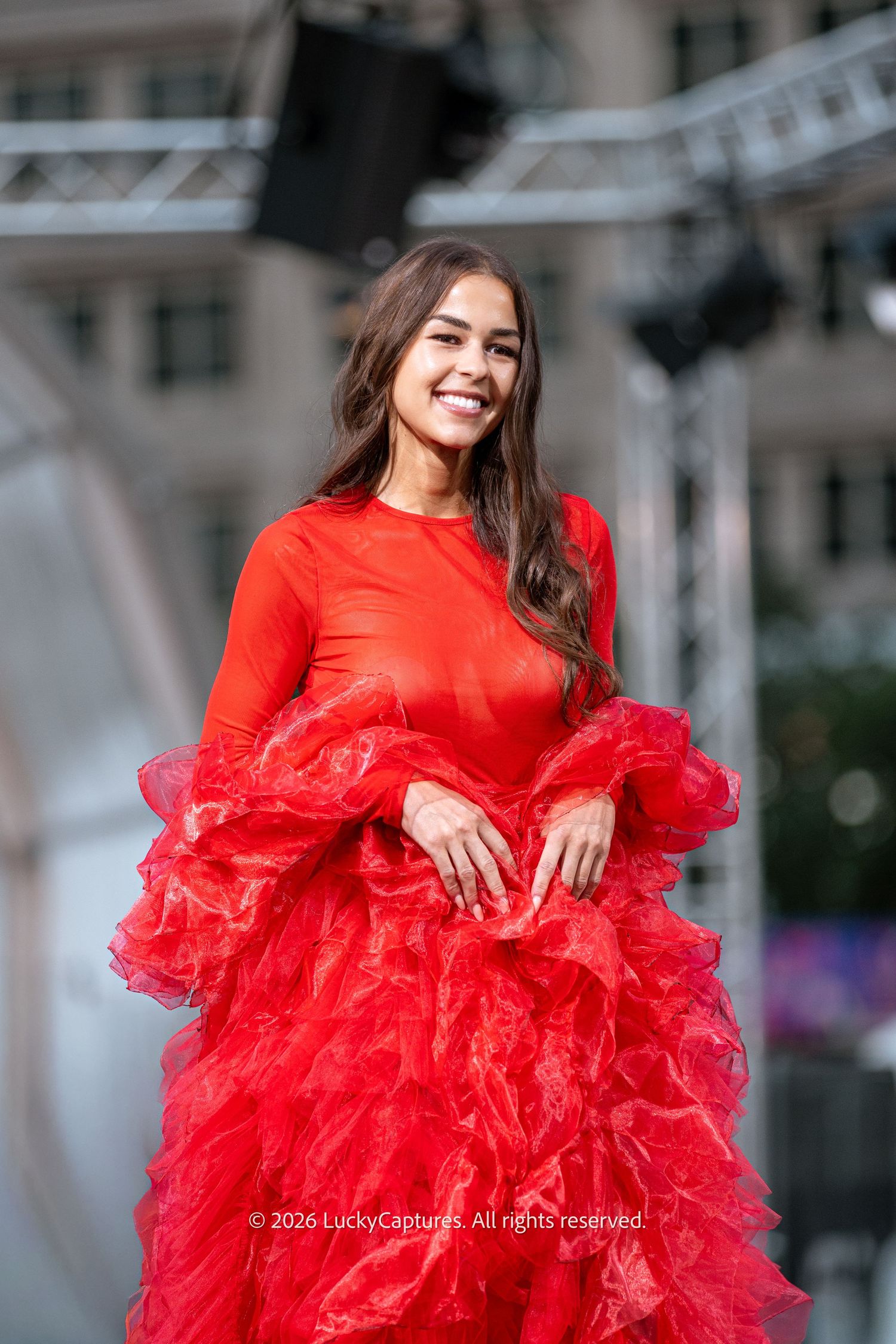 A smiling woman in a stunning red ruffled gown poses gracefully on an outdoor stage.