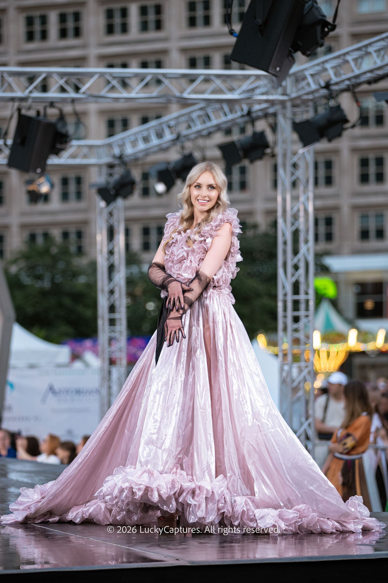 Elegant blonde model poses in pink ruffled ball gown with black lace gloves on outdoor runway.