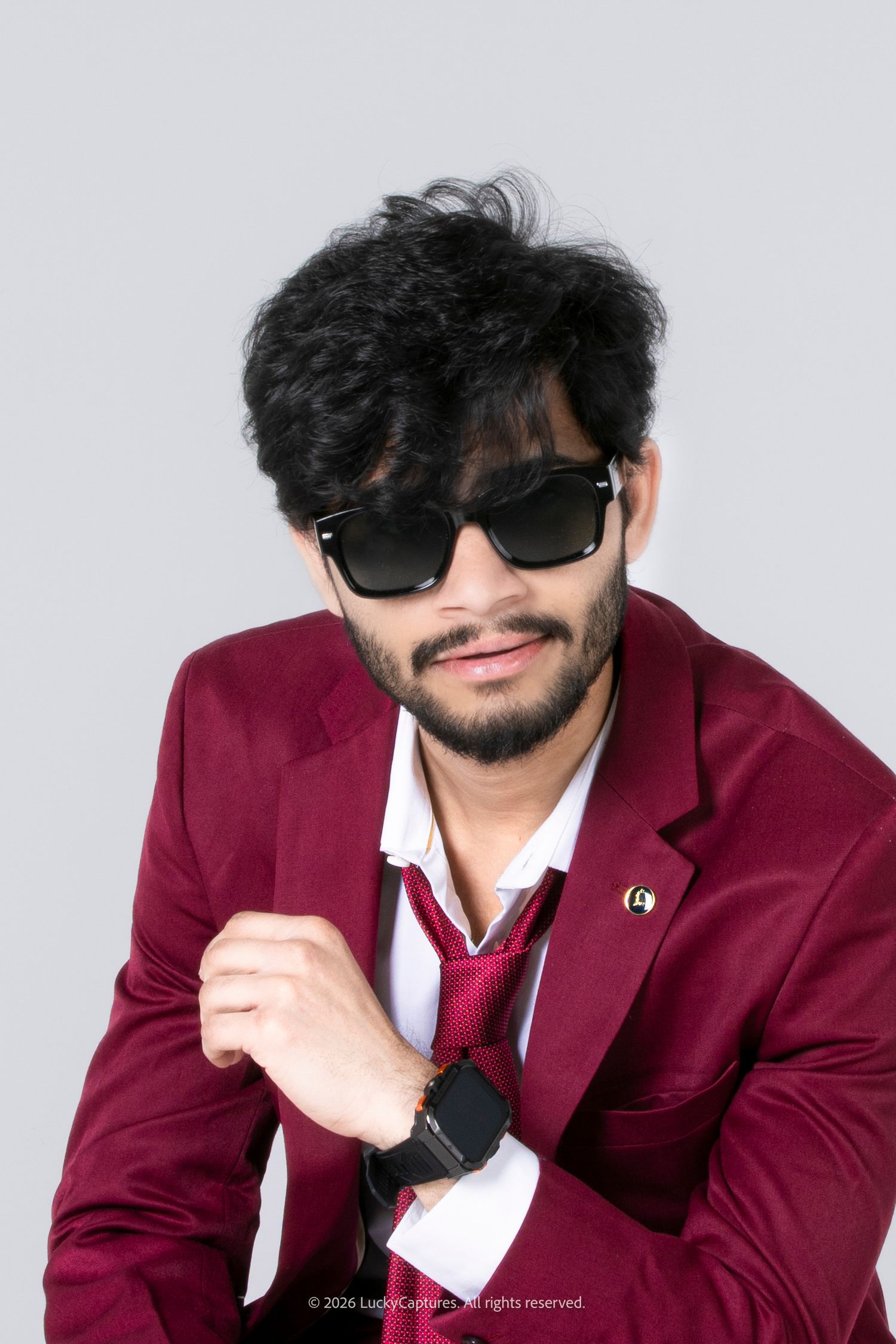 Young man in red blazer, white shirt, and sunglasses posing confidently with a watch and tie in a studio.