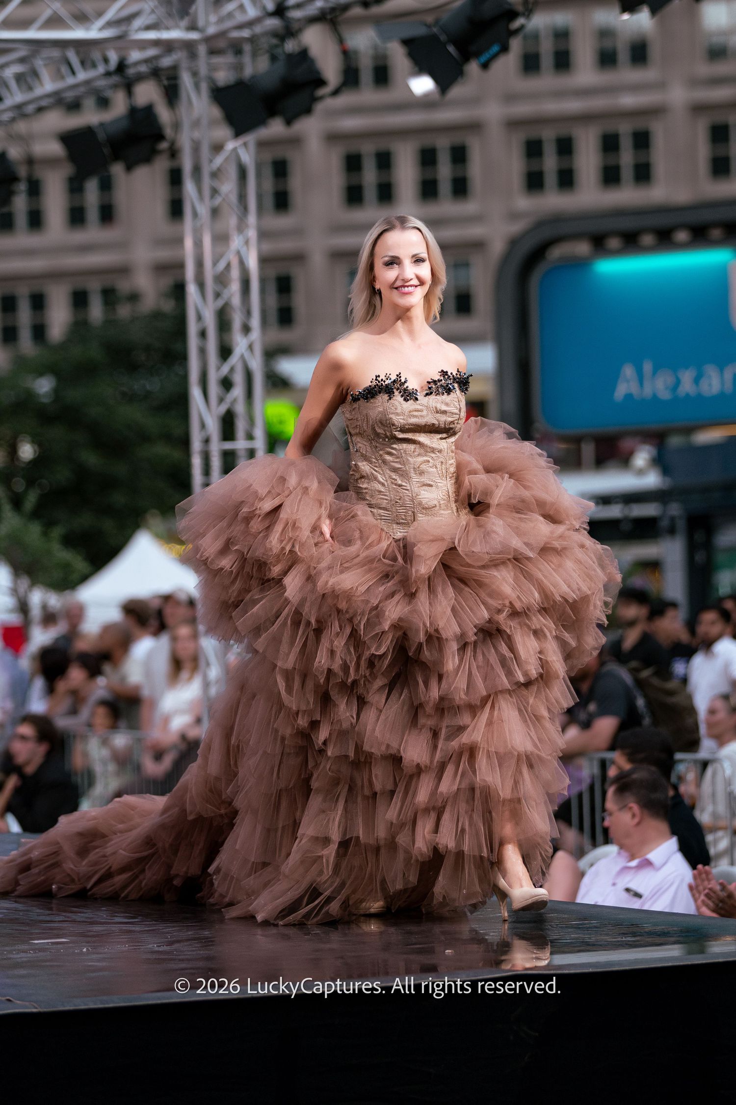 Smiling model struts outdoor fashion runway in dramatic brown layered tulle strapless ball gown.