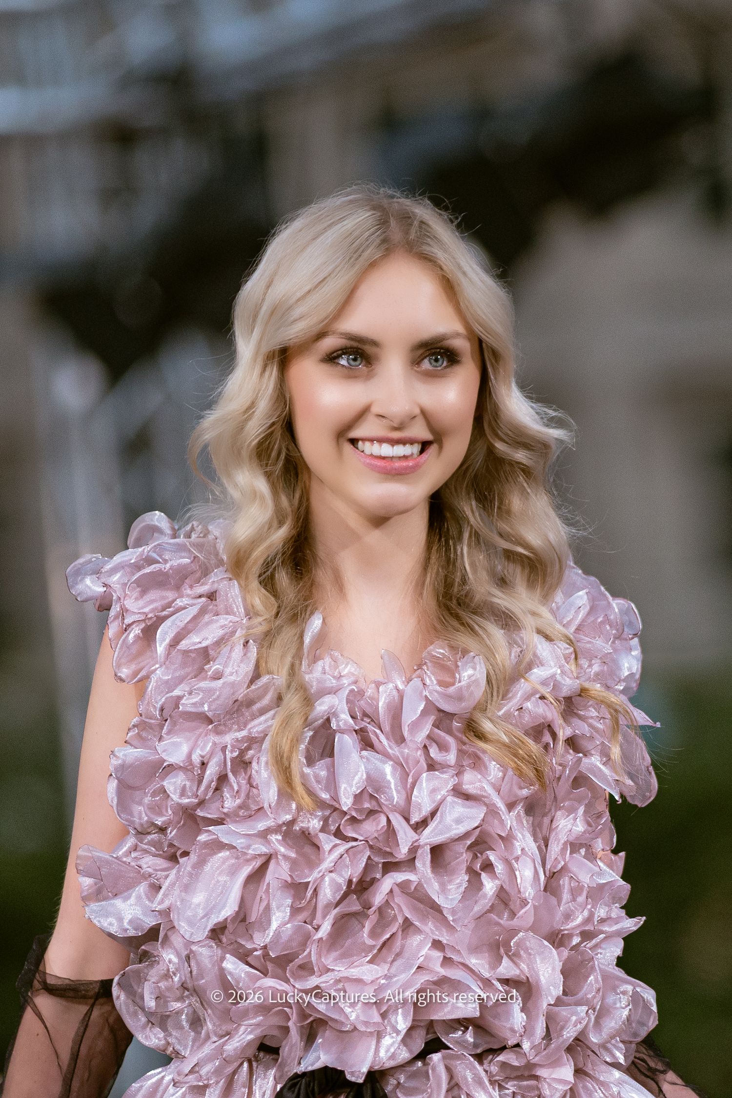 Close-up portrait of smiling blonde model wearing pink textured ruffle top at outdoor fashion event.