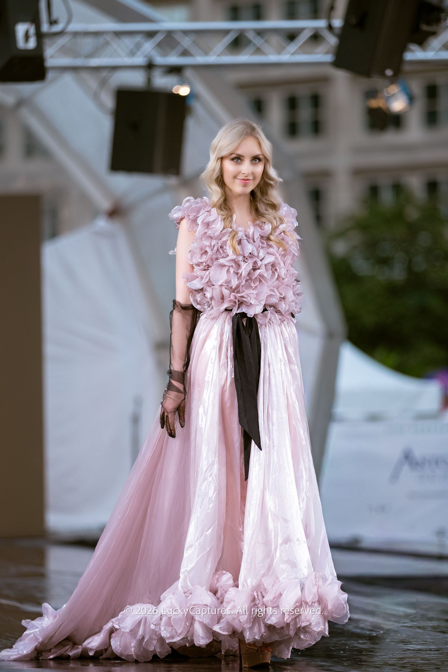 Blonde model showcases pink floral applique gown with black ribbon belt at outdoor fashion show.
