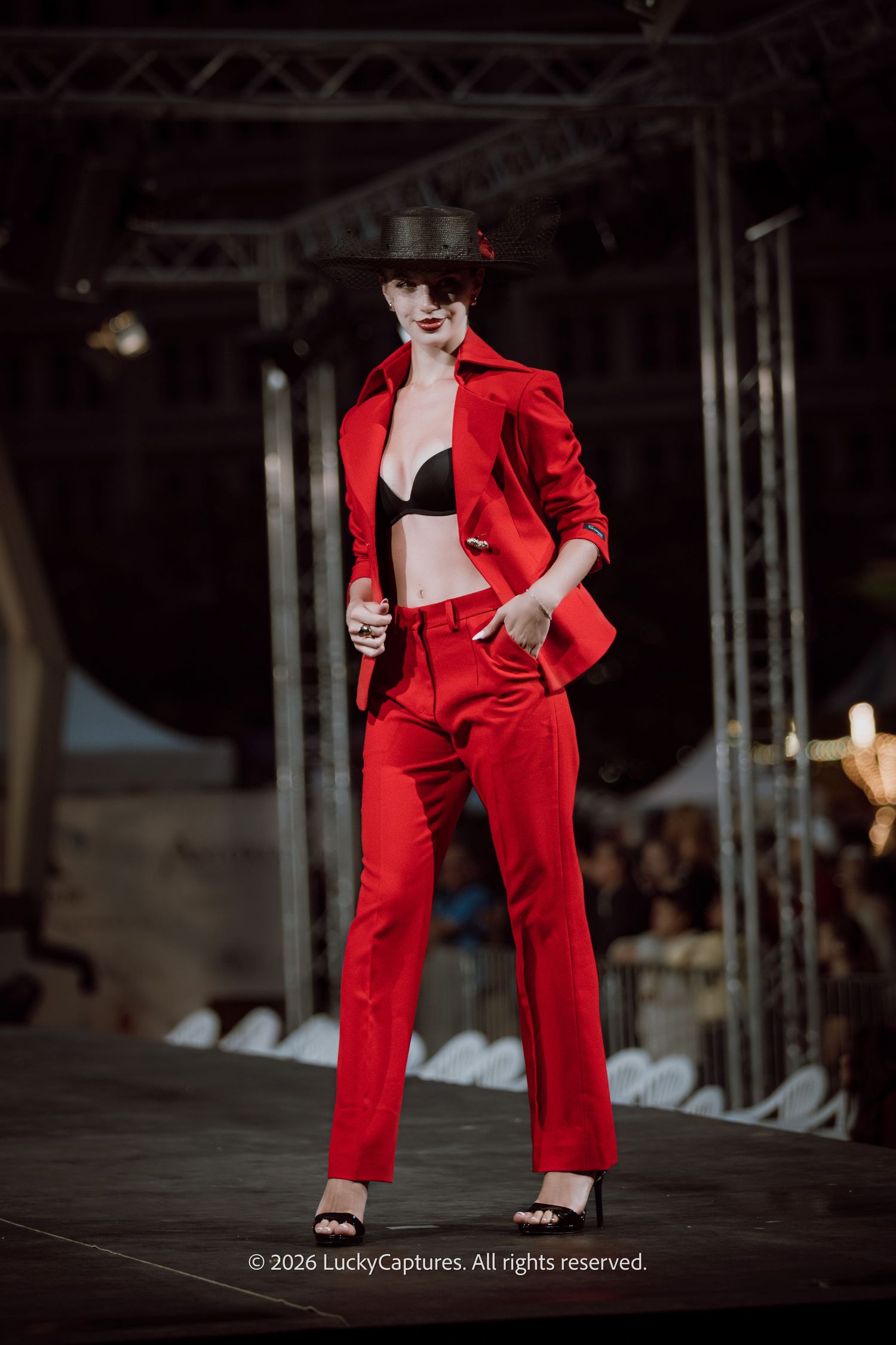 Model walks runway in red suit with black bra and wide-brim hat at a fashion show.