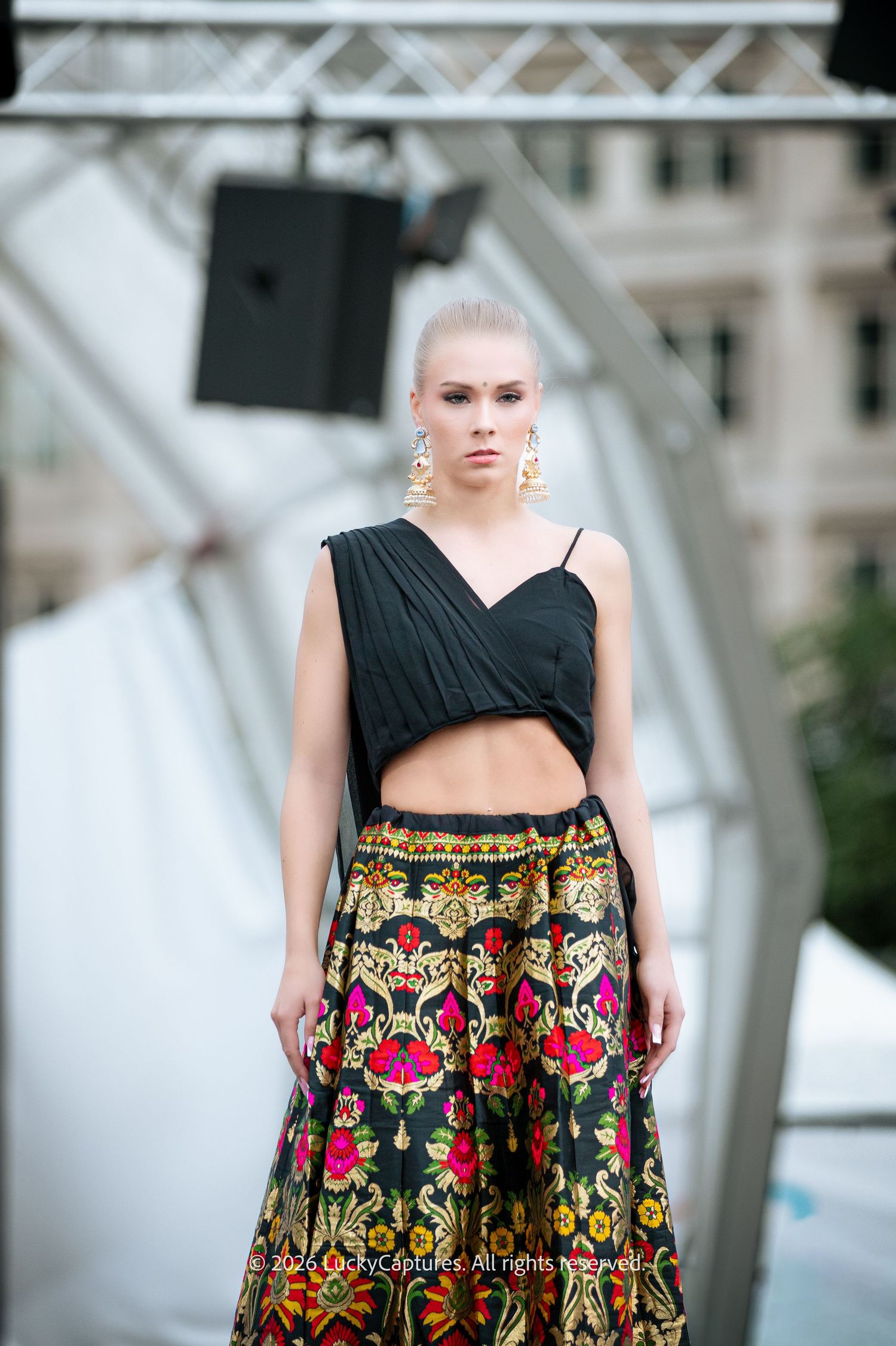Blonde model wearing a black crop top and colorful floral embroidered skirt poses outdoors at a fashion show.