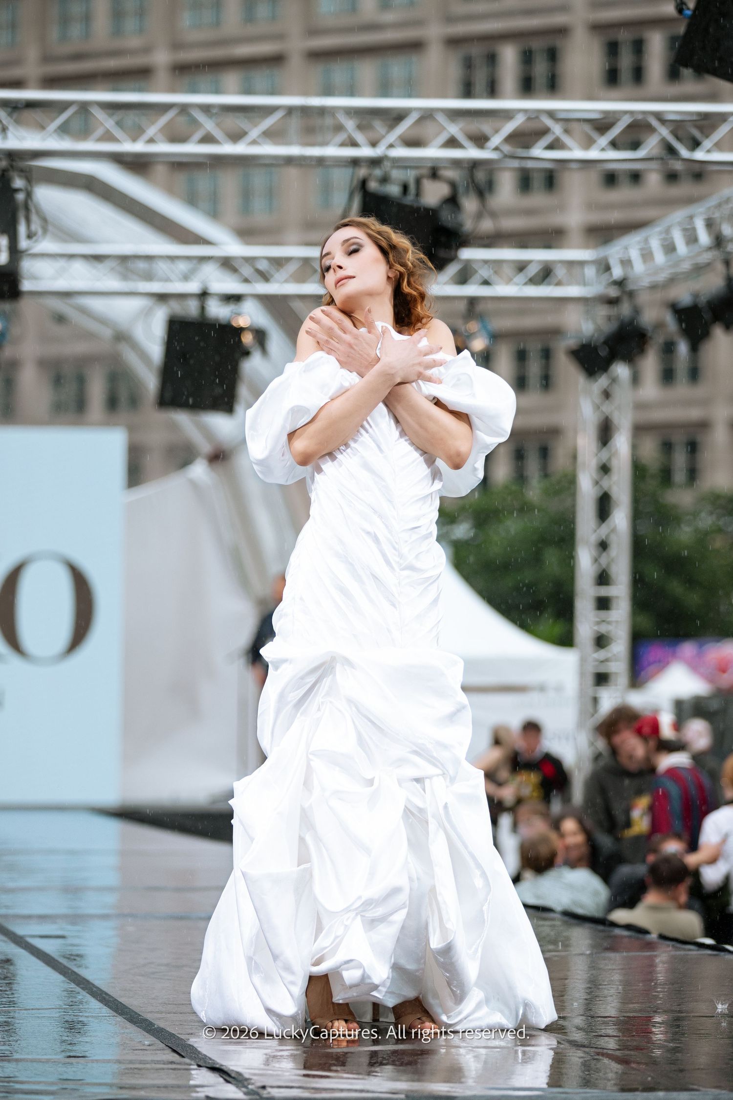 A woman in a white bridal gown poses gracefully on an outdoor runway stage during a fashion show.