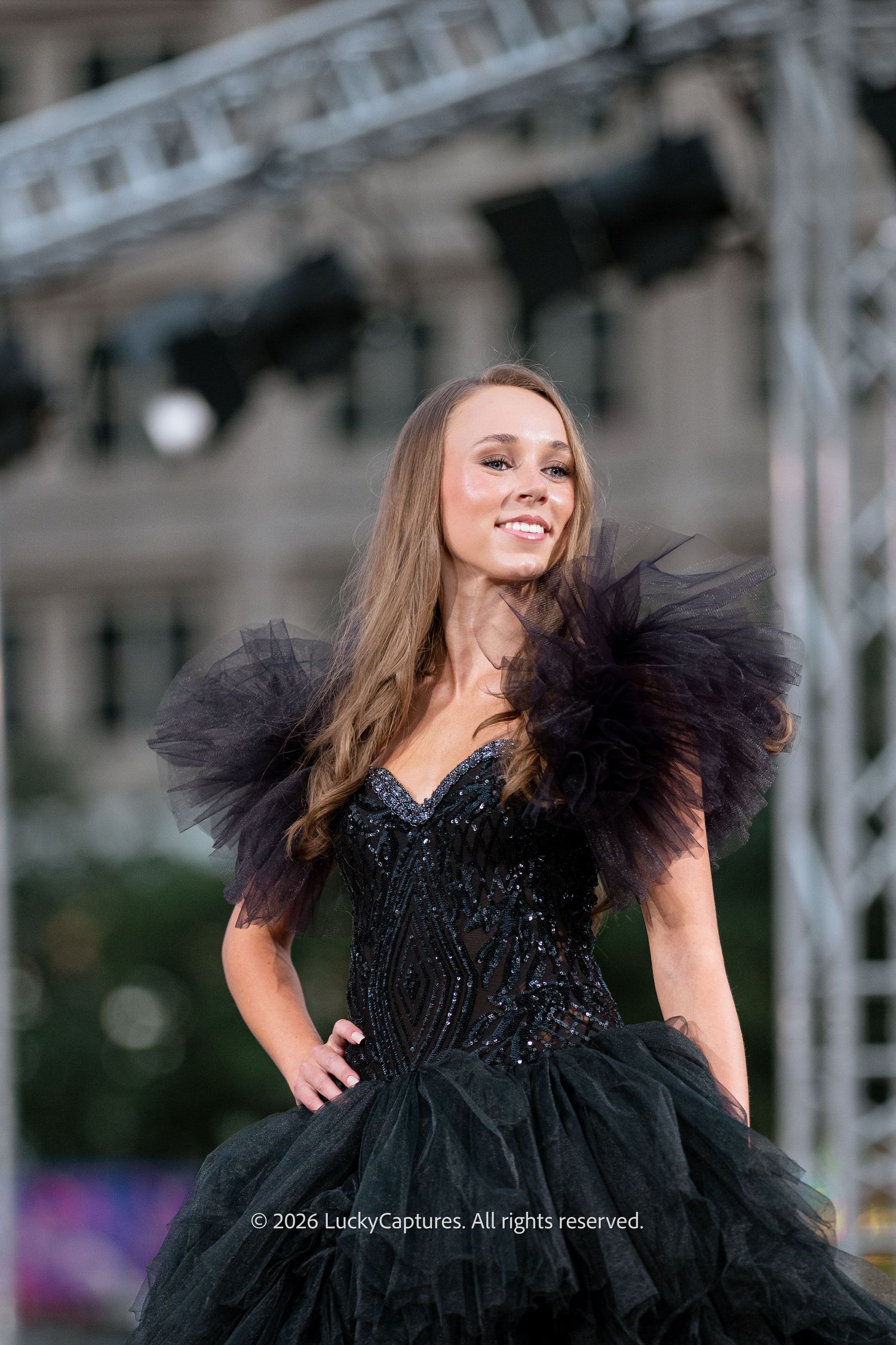 A young woman in a stunning black beaded corset ball gown with dramatic tulle ruffles poses on an outdoor stage.