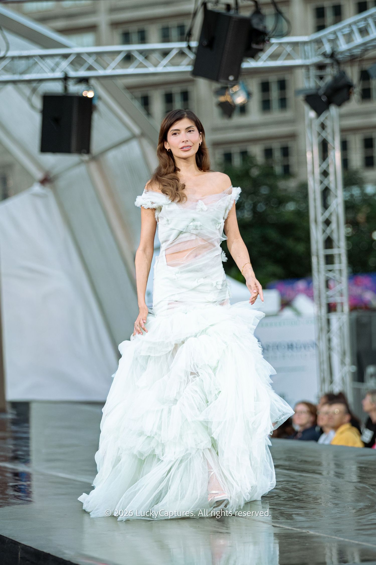 Model walks runway in sheer mint green off-shoulder ruffled mermaid gown at outdoor fashion show.