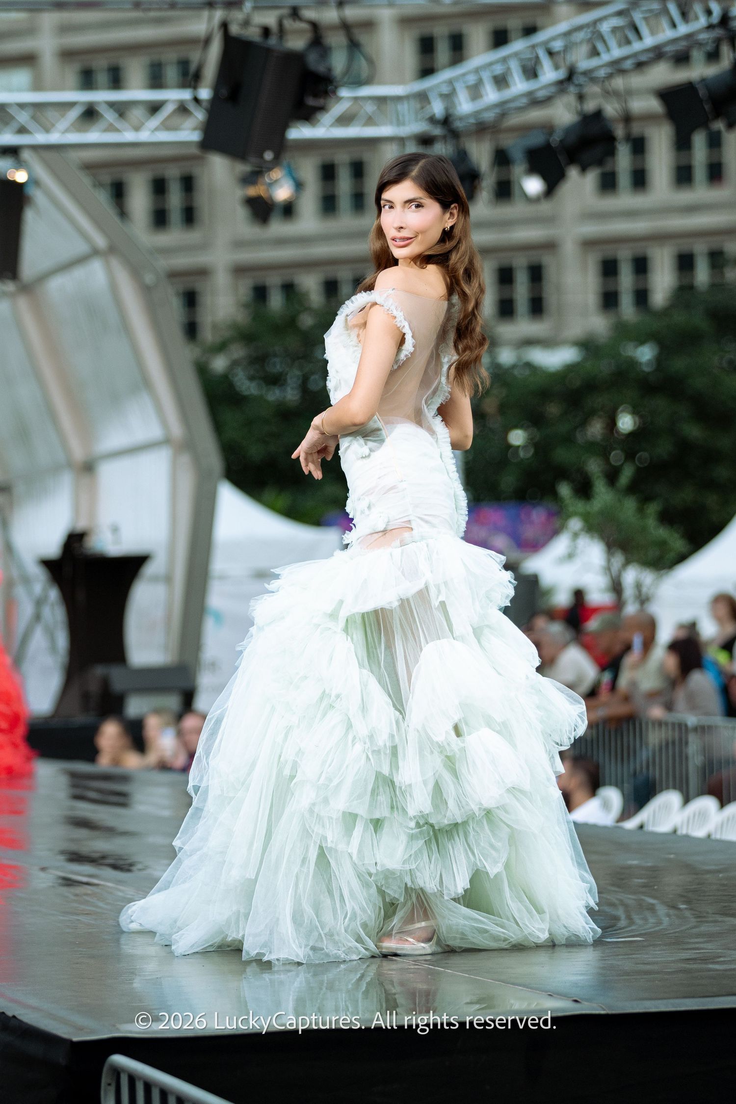 Model showcases backless mint green ruffled mermaid gown from behind on outdoor fashion runway.