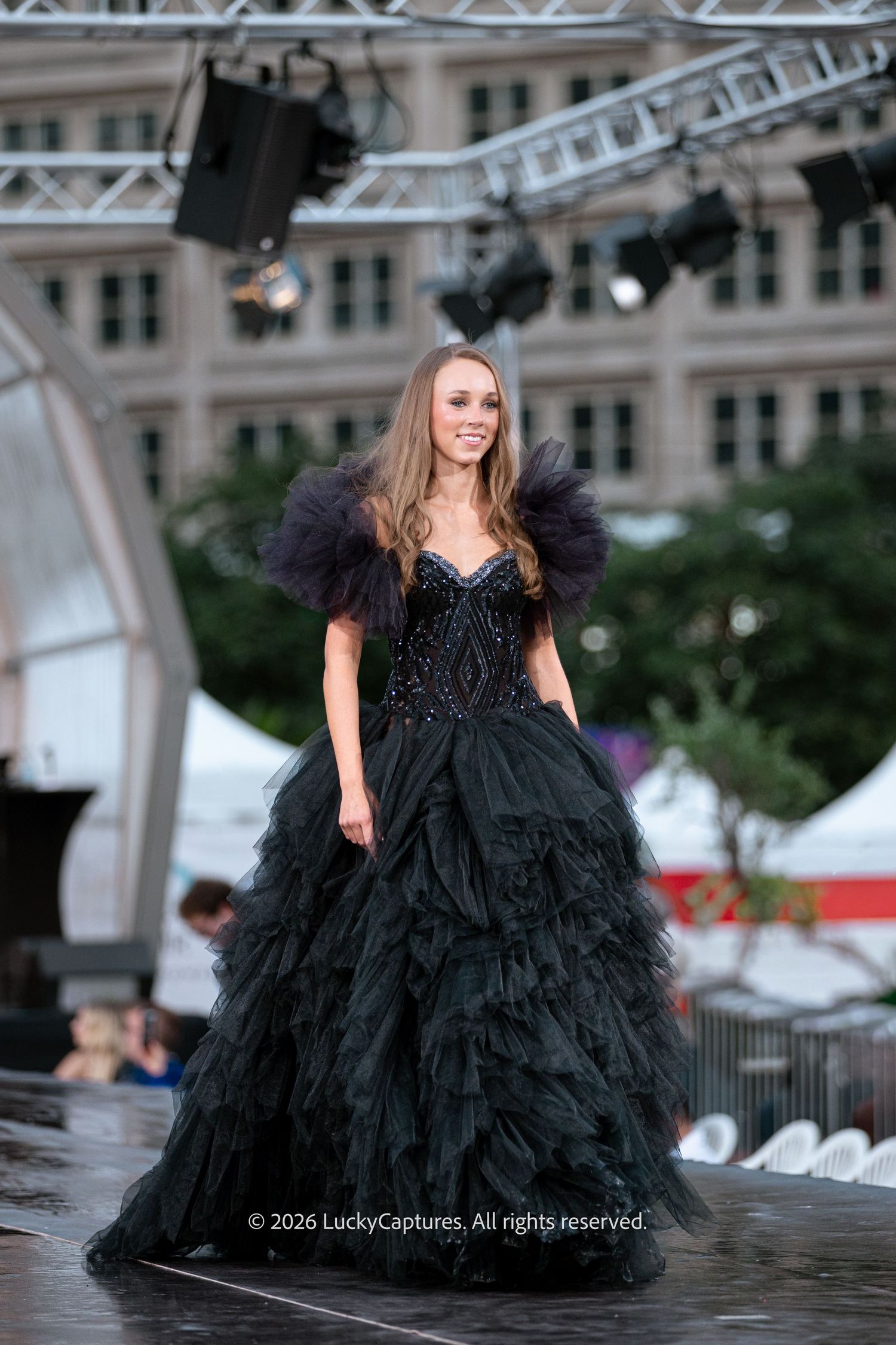 Model walks runway in stunning black tulle ball gown with ruffled shoulders at outdoor fashion show.