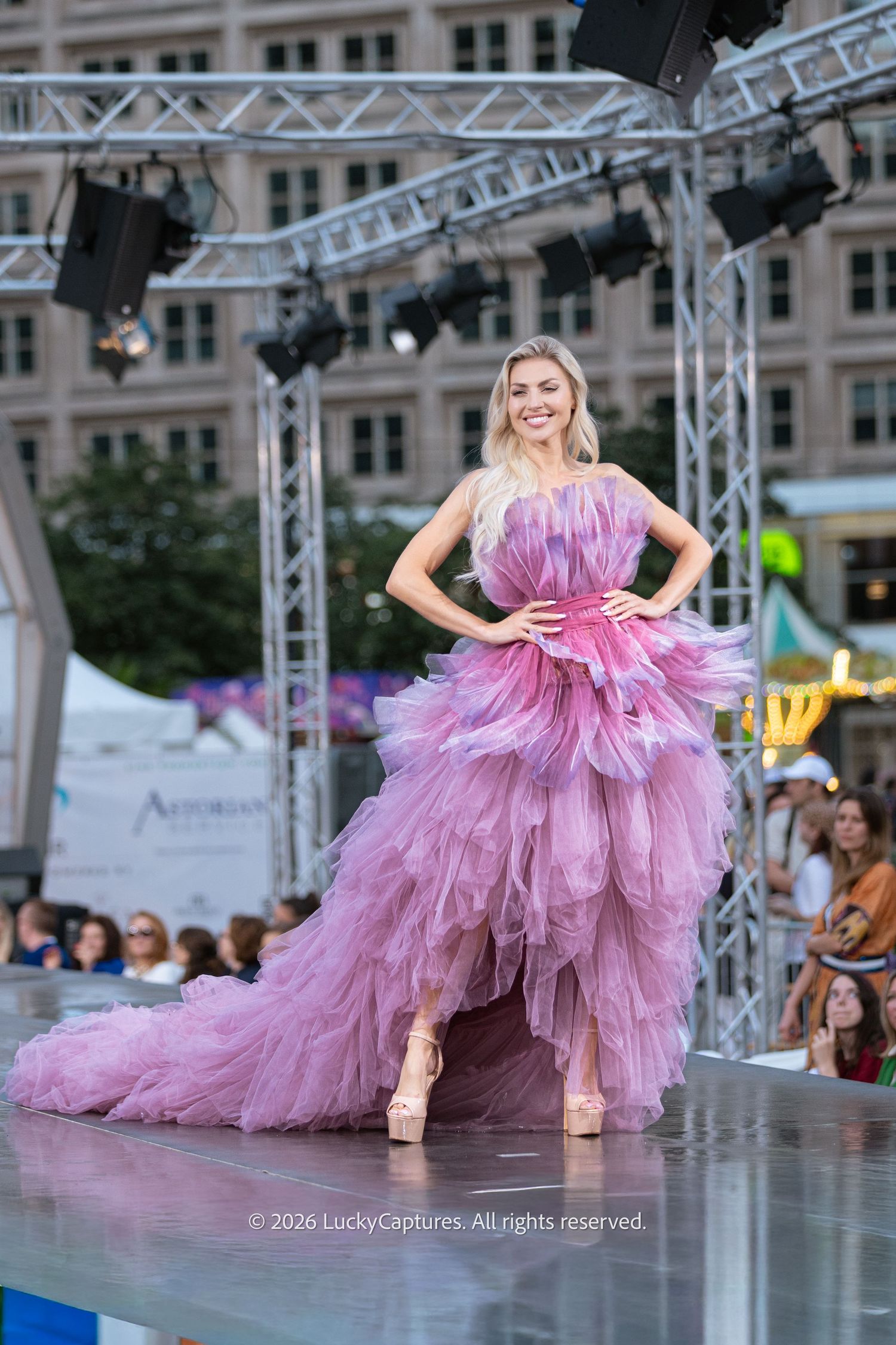 Confident blonde model poses hands-on-hips in pink layered tulle high-low gown on outdoor fashion runway.