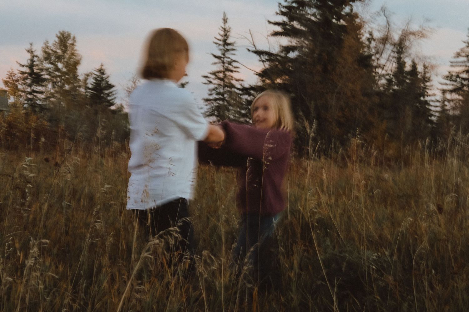 Two children playfully spinning together in a golden autumn field surrounded by tall grass and trees.