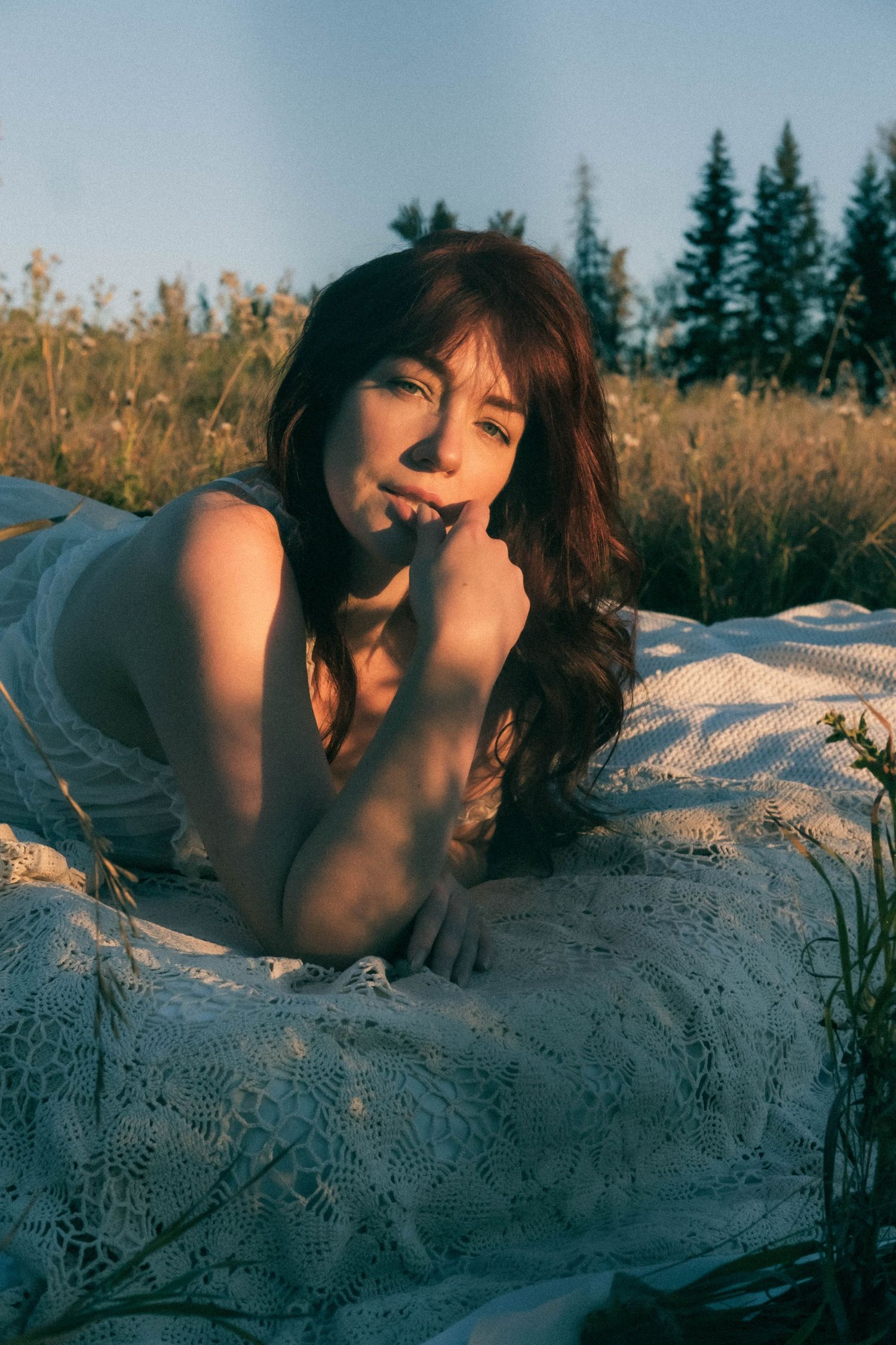 A young woman with long red hair lies on a white lace blanket in a sunlit meadow, gazing thoughtfully at the camera.