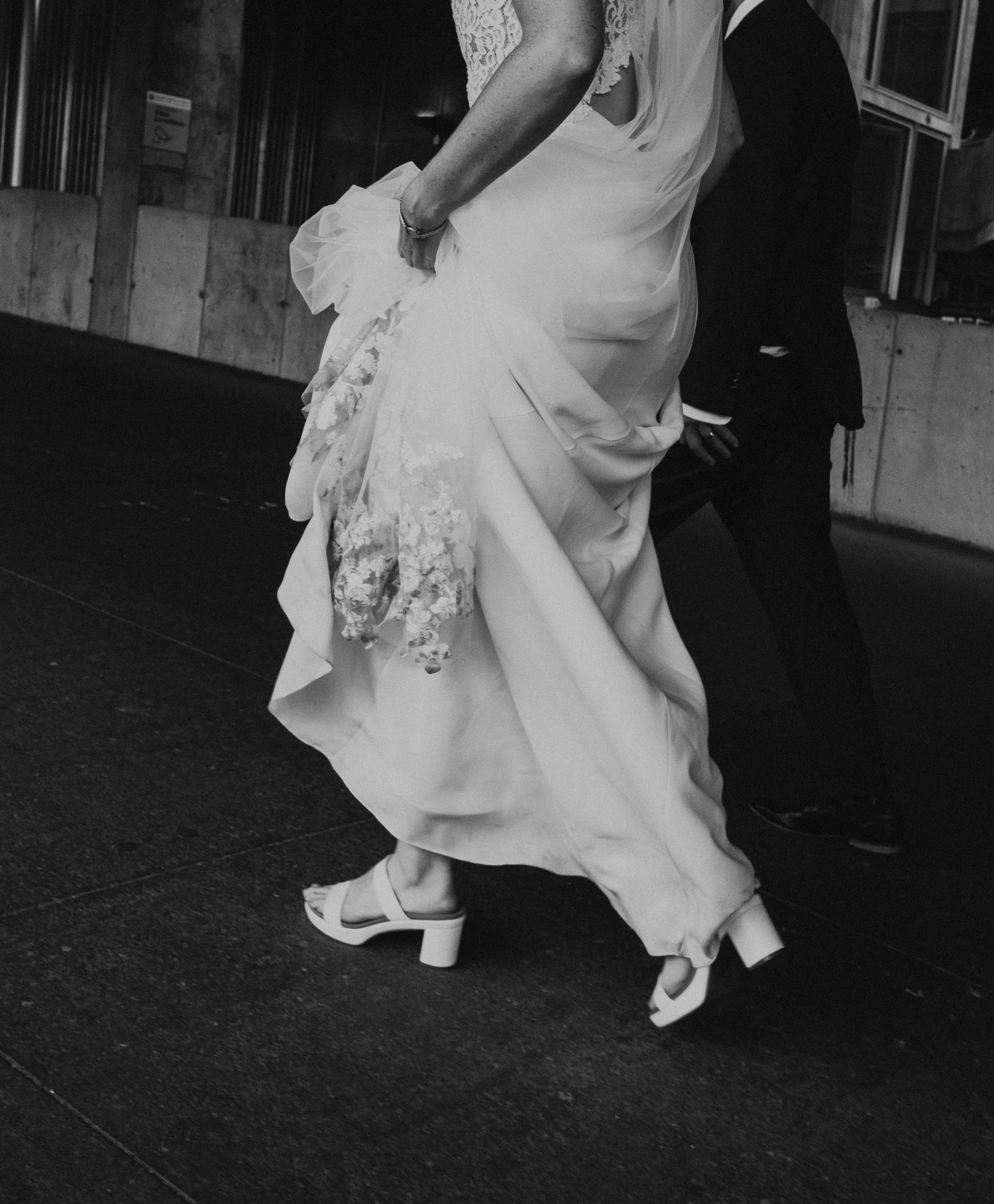 Black and white photo of a bride in a lace wedding gown and heels walking with a groom in a suit.