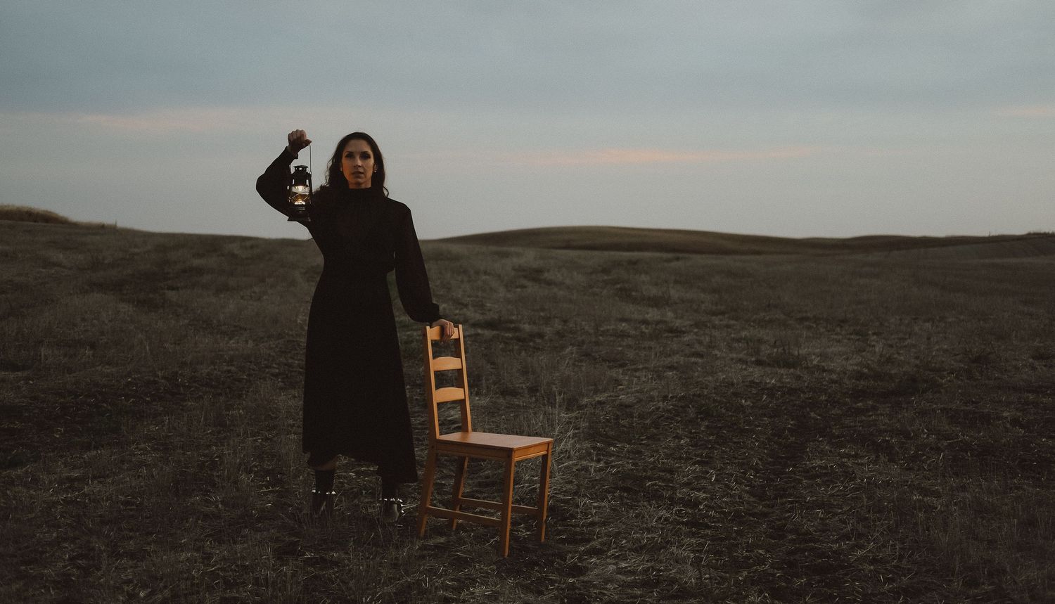 A woman in a black dress holds a lantern while standing beside a wooden chair in a vast open field.