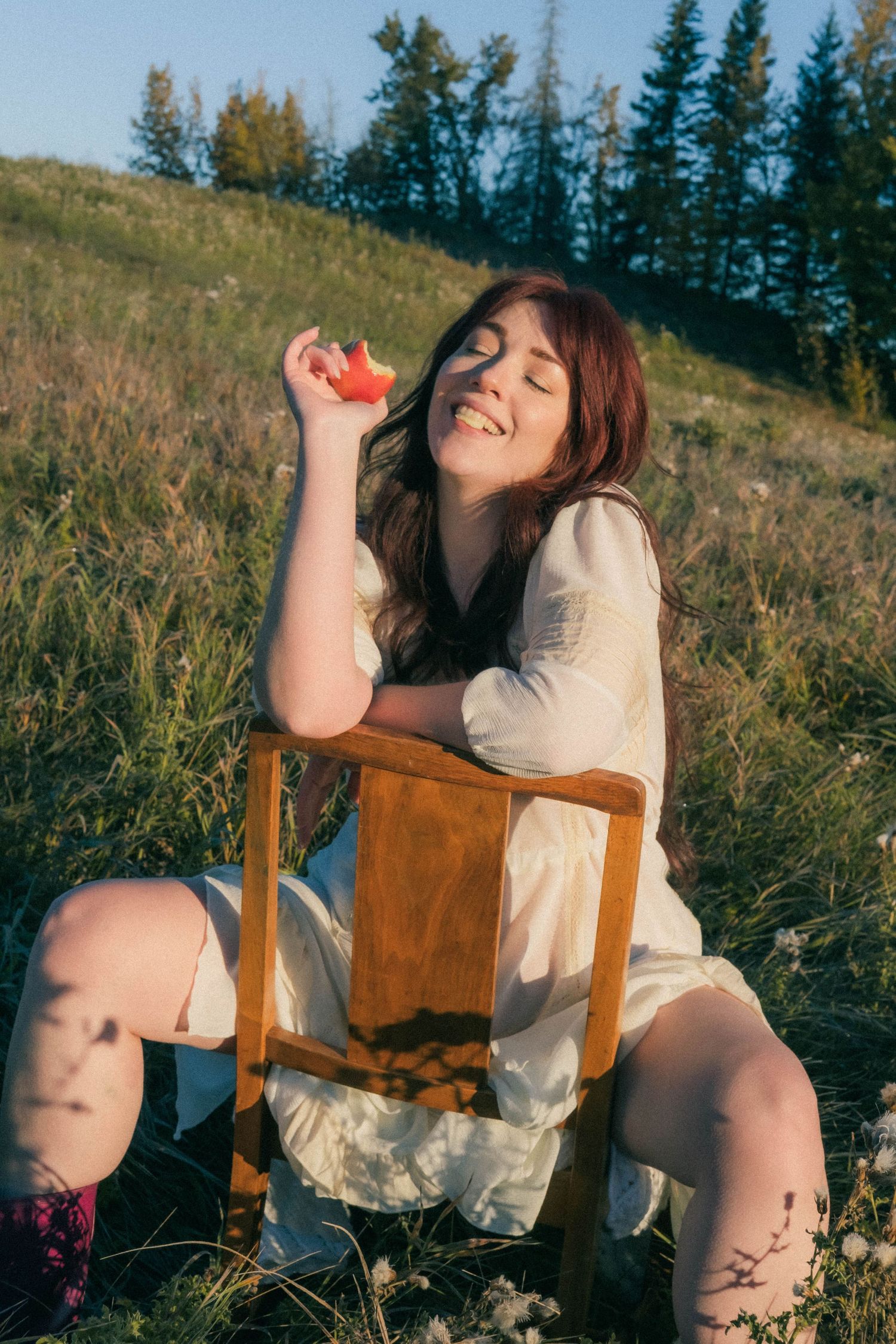 Young woman in white dress sitting on wooden chair in sunlit meadow, smiling with eyes closed, holding red flower.